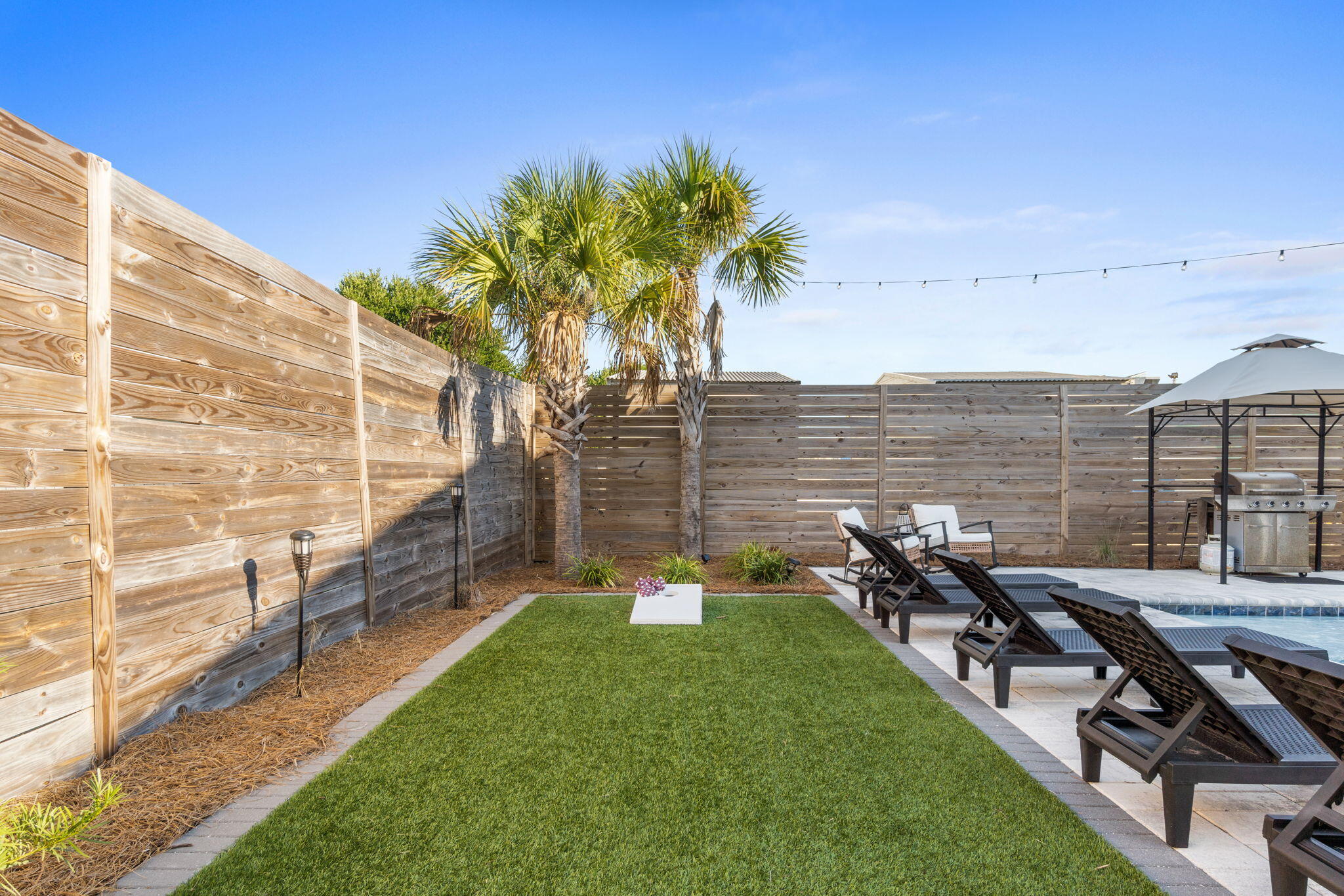 Fenced backyard with lawn, palm trees, poolside loungers, and a covered grill area.