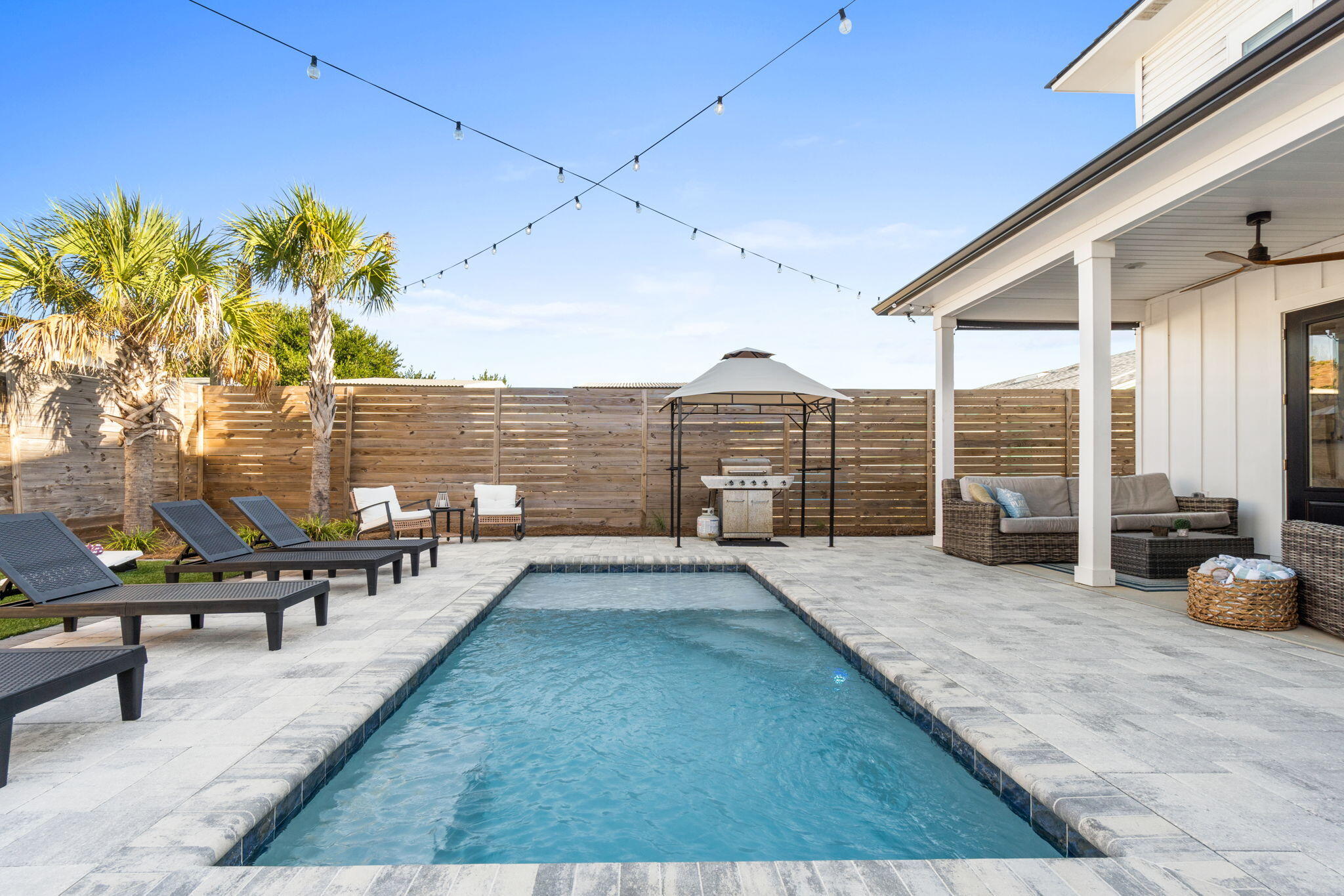 Backyard pool beside a covered patio with lounge chairs, palm trees, and a barbecue grill.