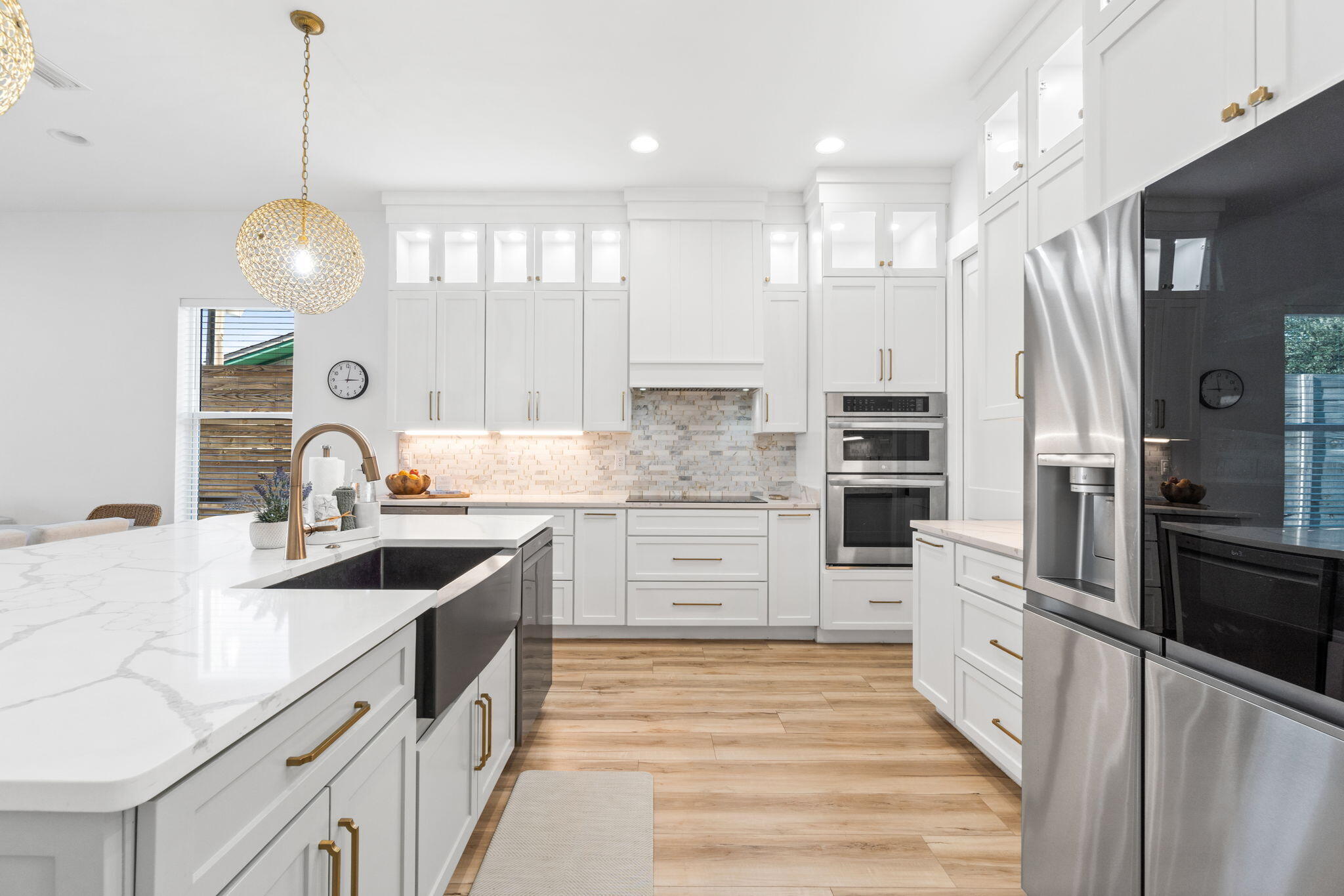 Bright white kitchen with a large marble-look island, brass faucet, stainless steel appliances, and wood flooring