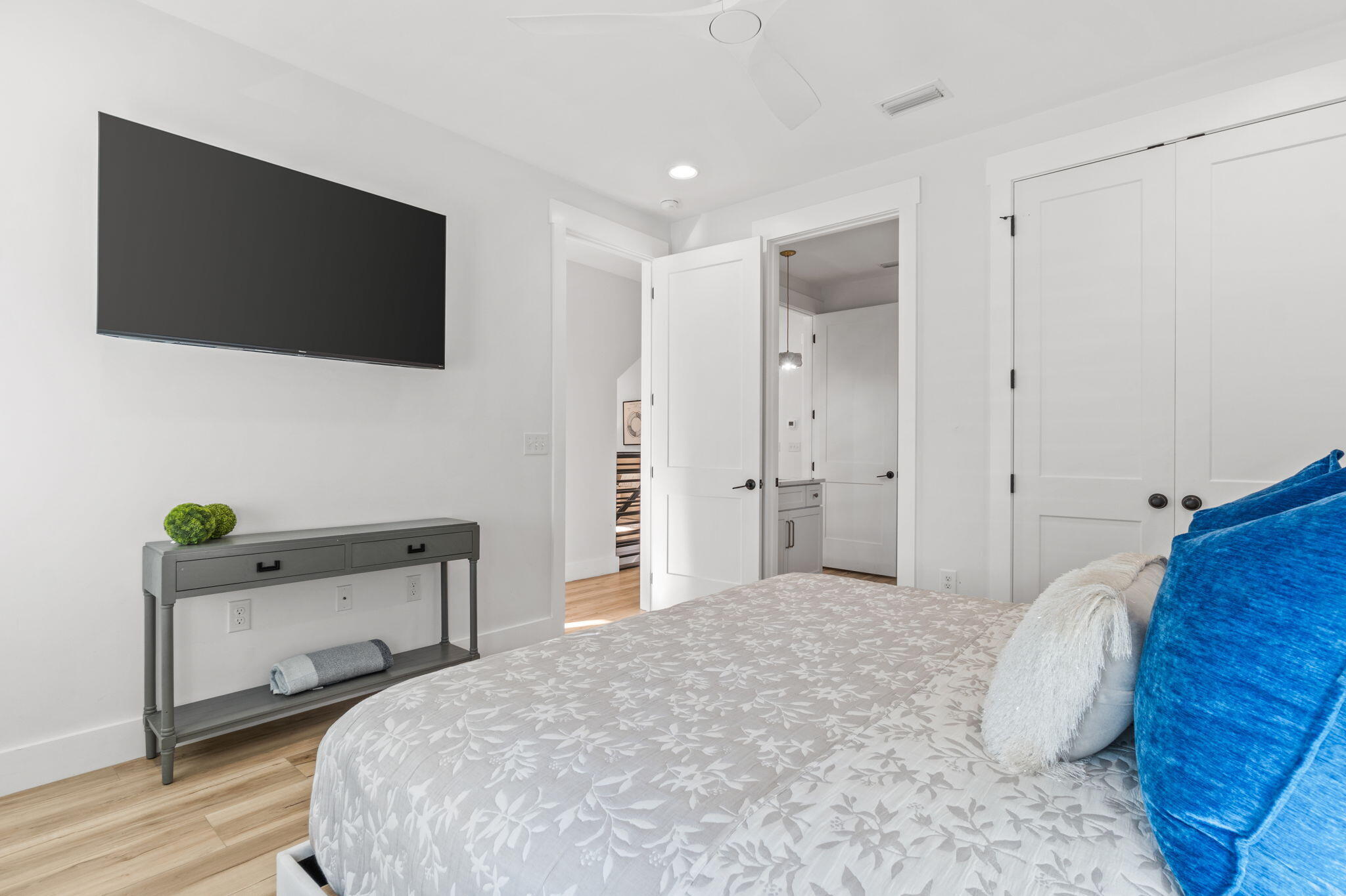 Furnished bedroom with a floral quilt, bright blue pillows, wall-mounted television, and open bathroom doors.