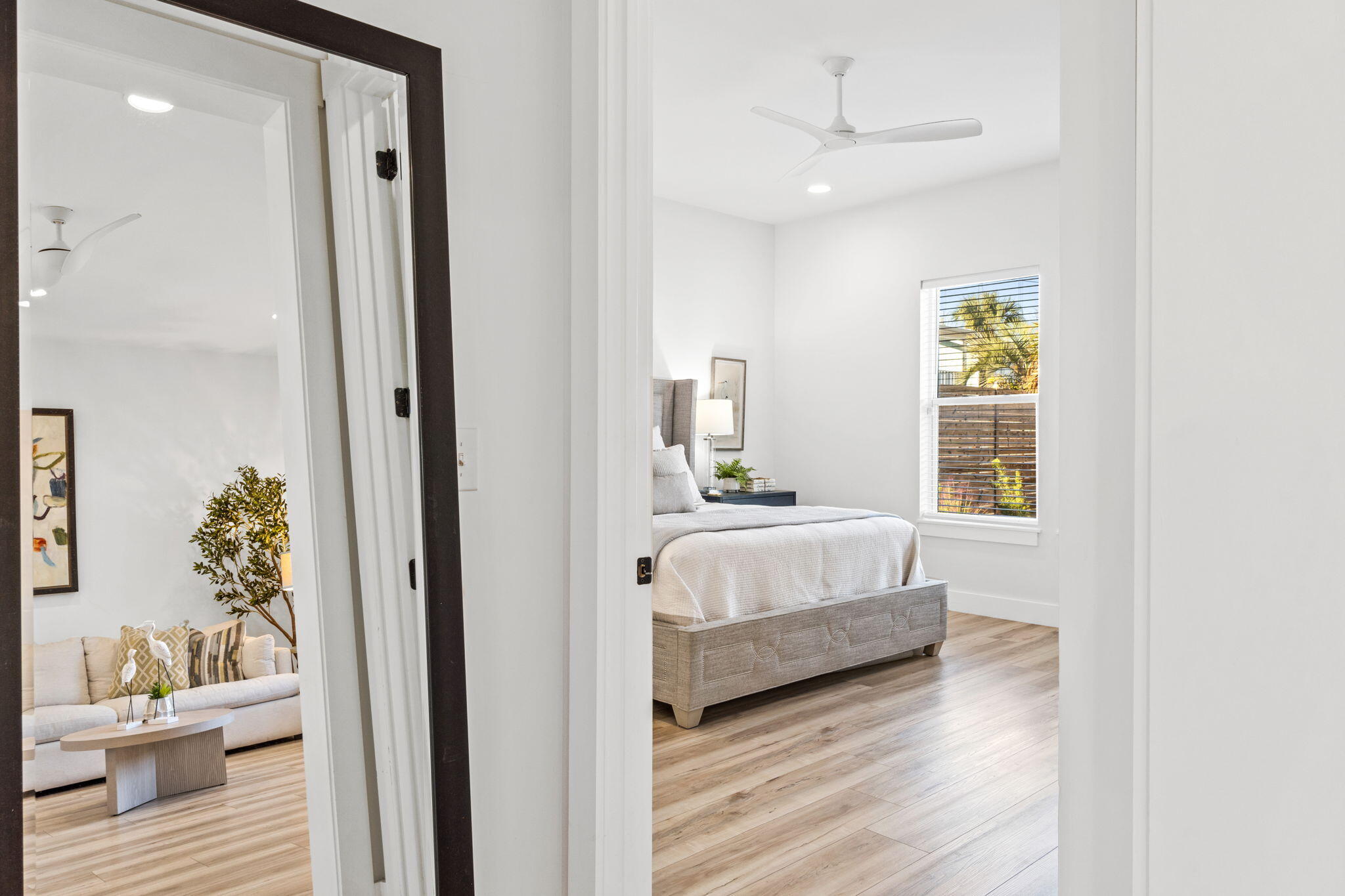 Staged bedroom with a gray upholstered bed, wood-look flooring, and a window overlooking palm trees.