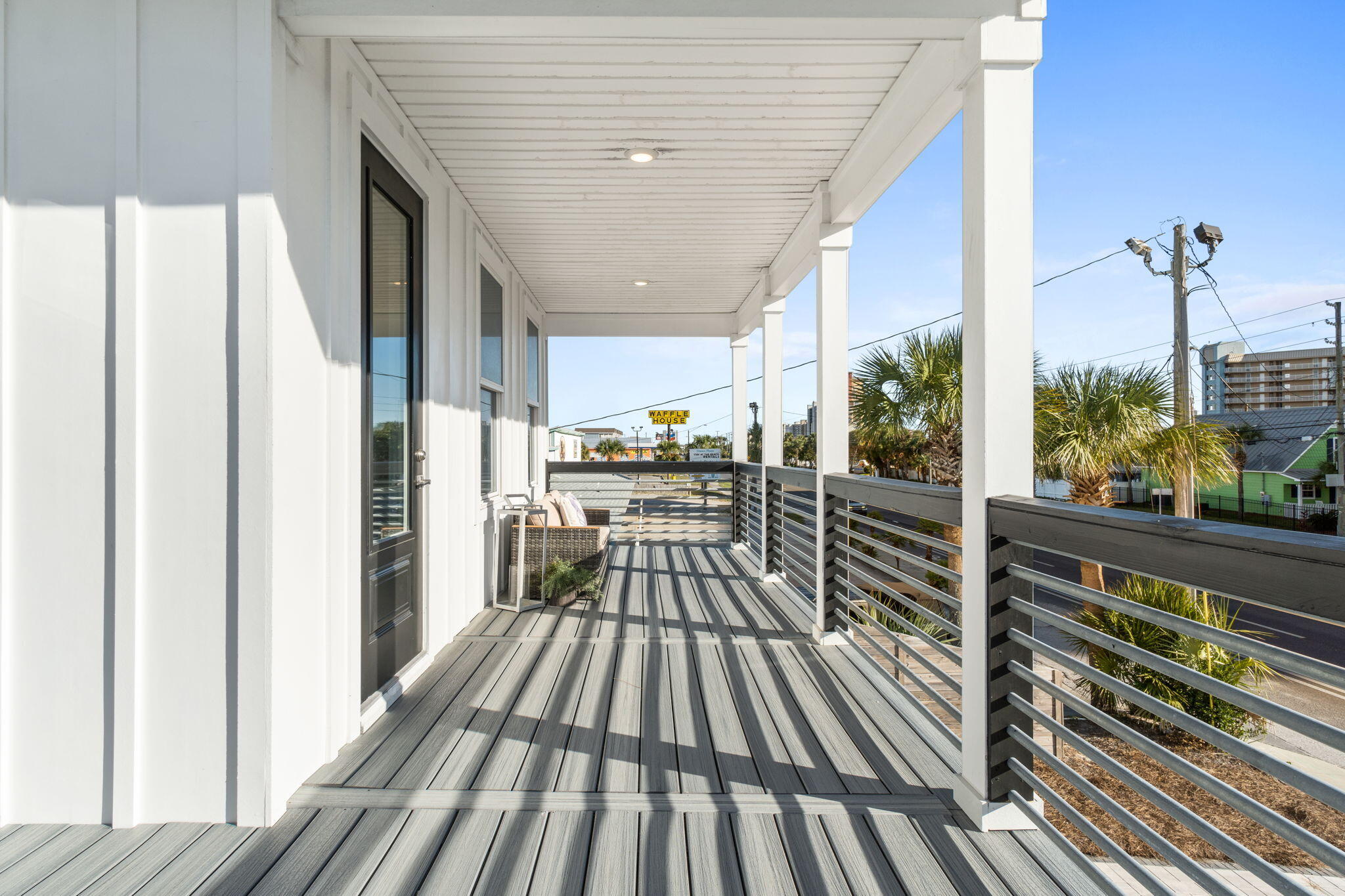 Covered balcony with gray decking, dark railings, and a small seating area overlooking palm trees and nearby buildings.