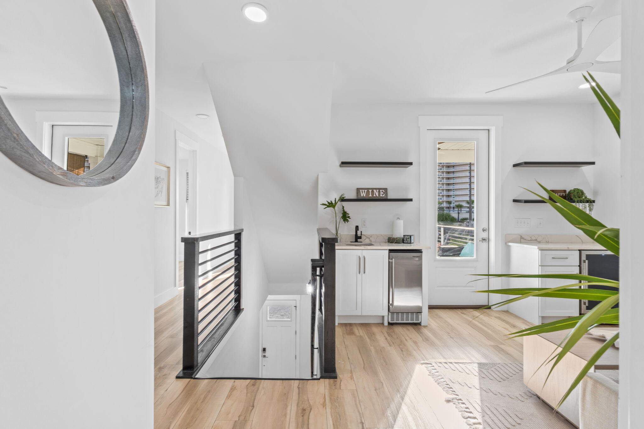Bright condo kitchenette with white cabinets, beverage refrigerator, floating shelves, and glass balcony door.