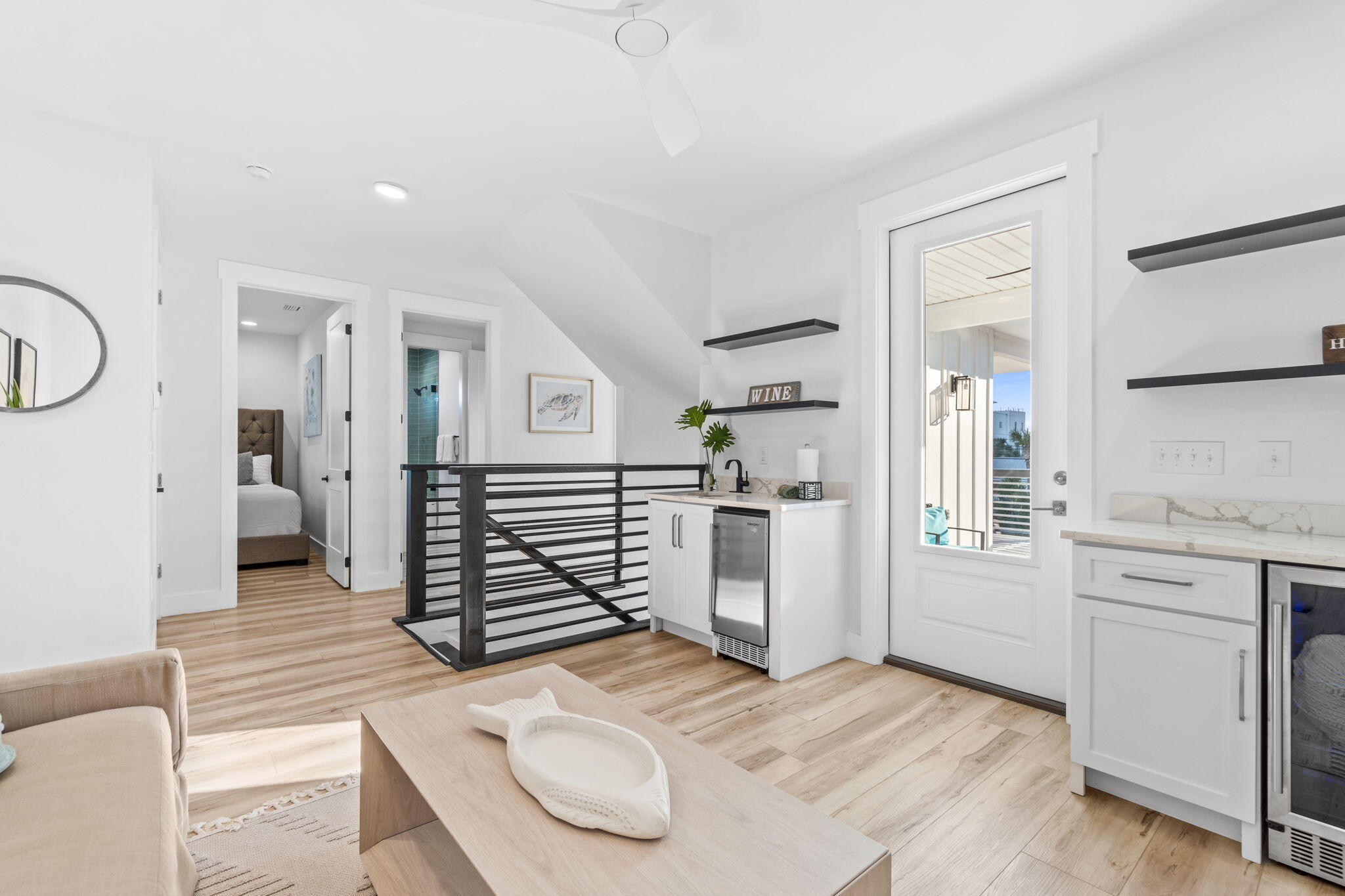Bright living area with compact kitchenette, black stair railing, and glass-paneled balcony door.