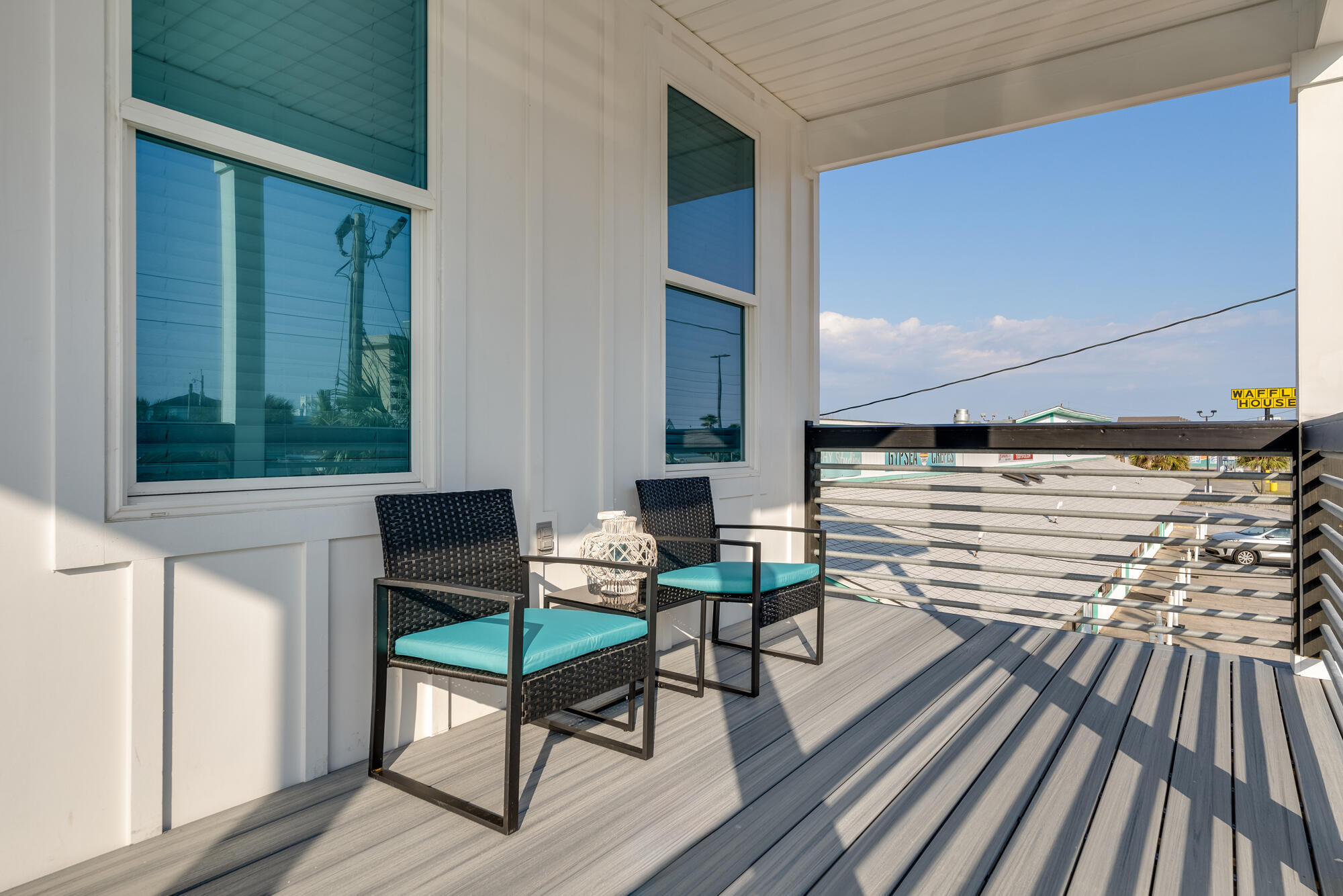 Furnished balcony with turquoise-cushioned chairs overlooking nearby shops and a parking lot.