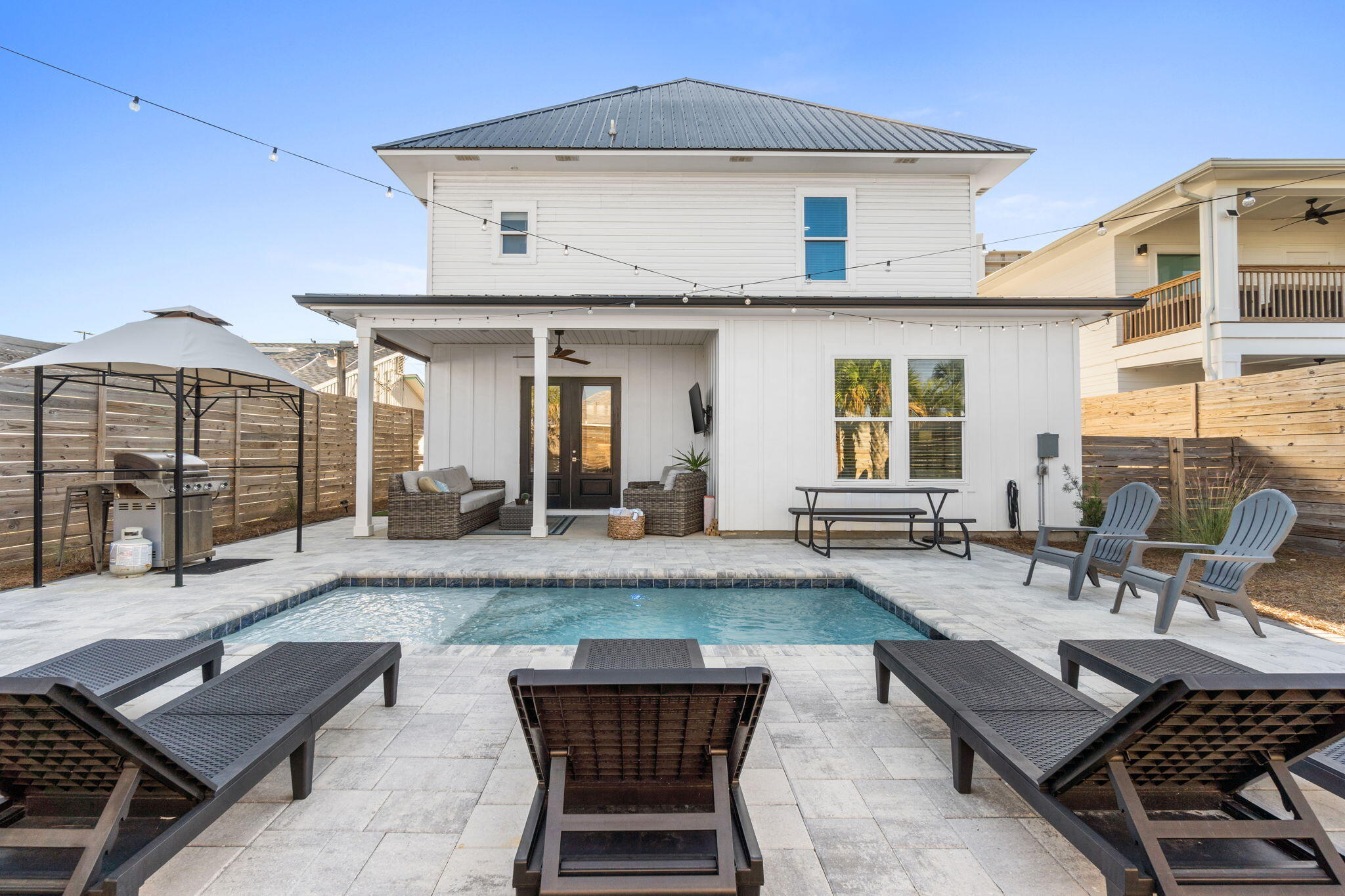 Backyard pool and patio with lounge chairs and a covered seating area beside a white two-story home.
