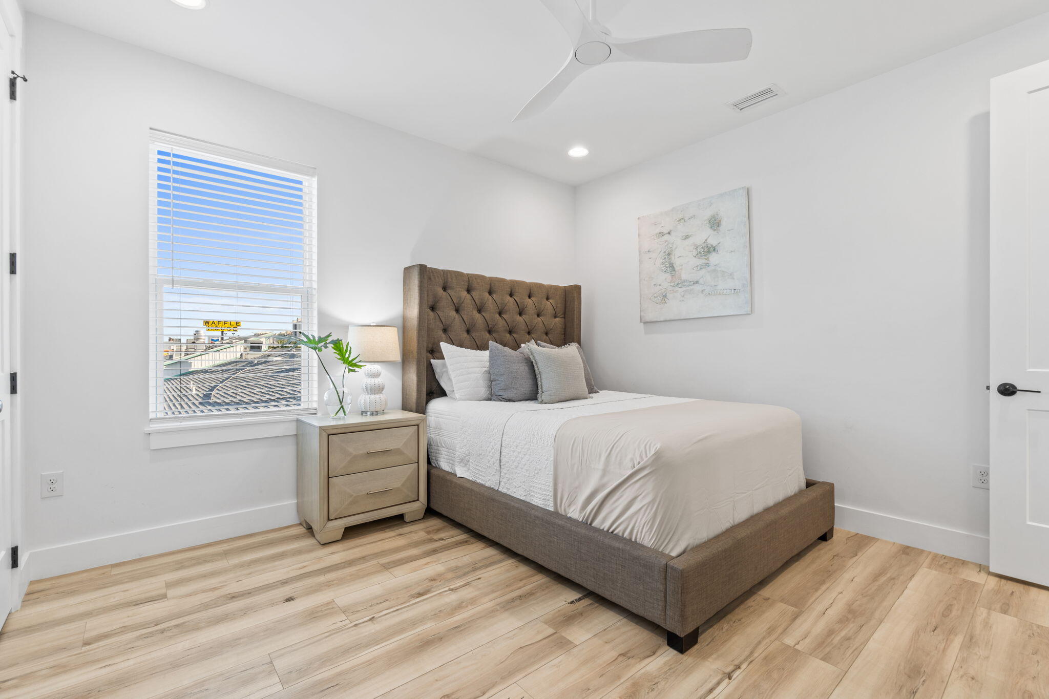 Furnished bedroom with a brown upholstered bed, white bedding, and a window overlooking rooftops.