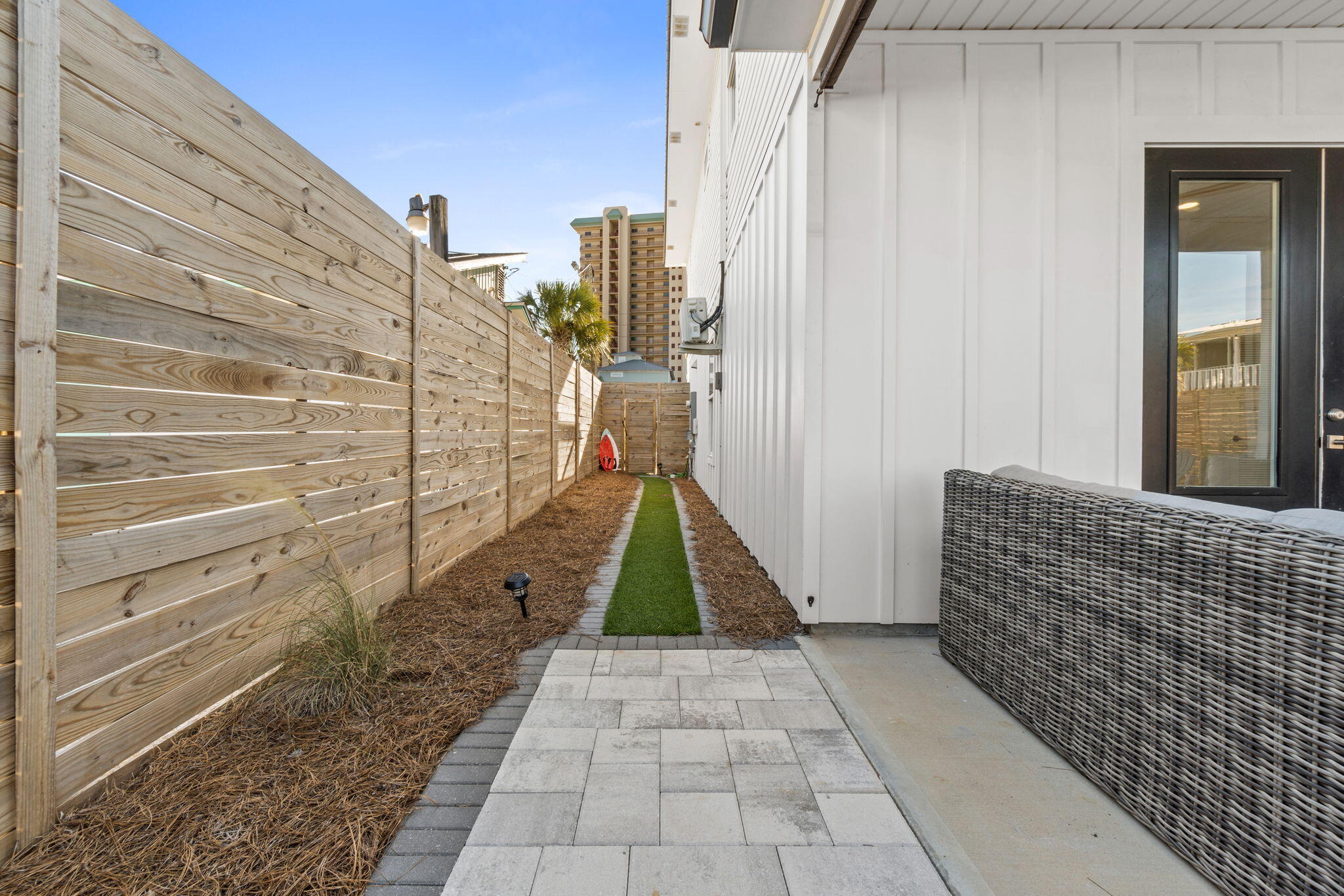 Side yard with a paved walkway, wooden privacy fence, artificial grass strip, and wicker patio seating.