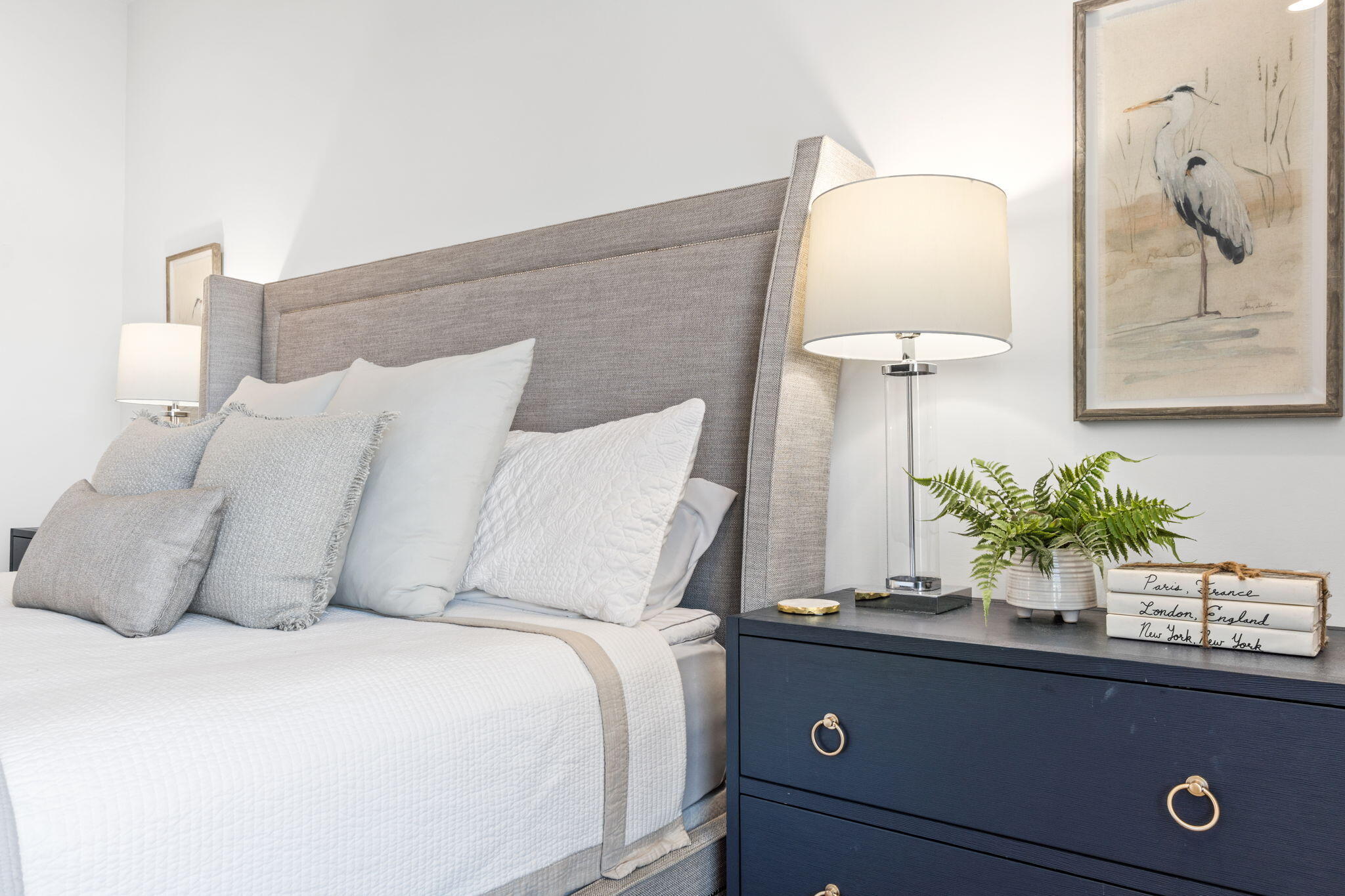 Staged bedroom with a white quilted bed, tall gray upholstered headboard, and navy dresser.