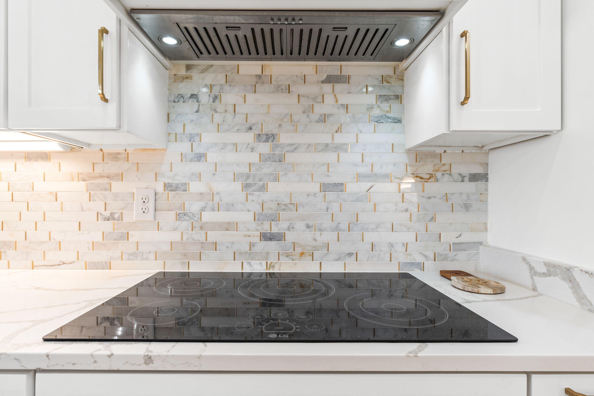 Kitchen cooktop beneath a stainless hood, framed by white cabinets and a veined marble backsplash.