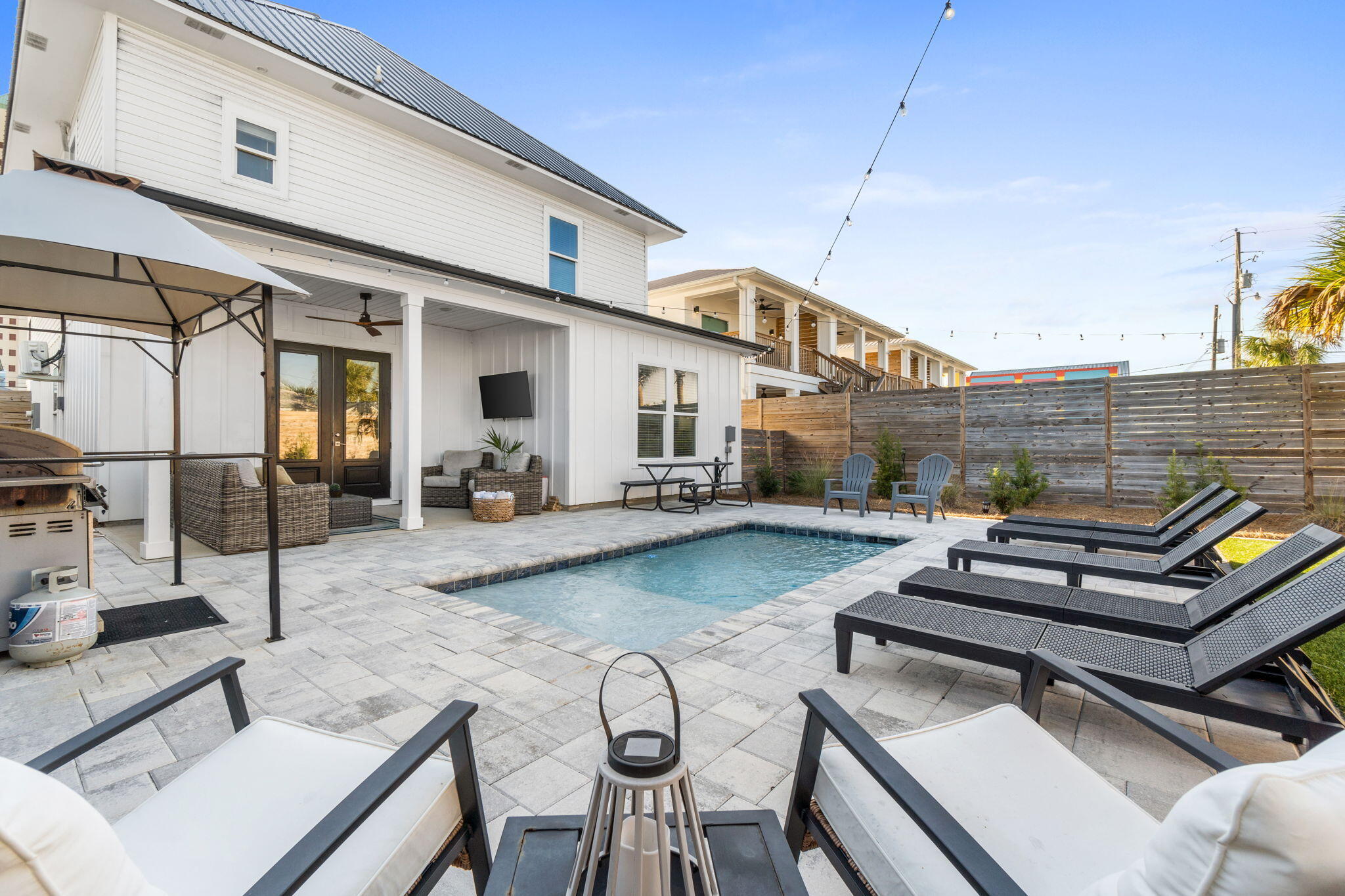Backyard pool patio with black loungers, outdoor seating, and string lights beside a white home.