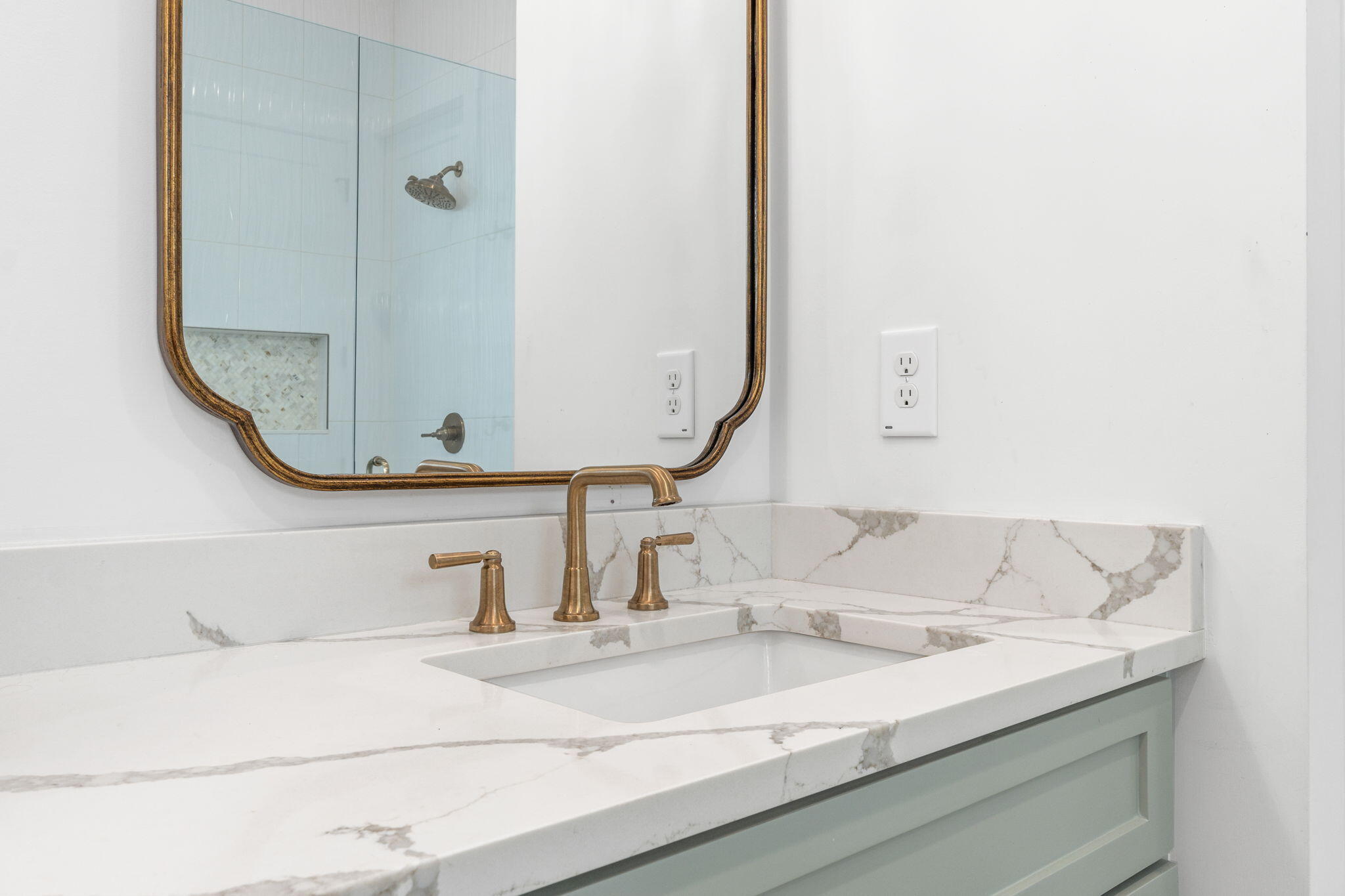 Bathroom vanity with marble-look counters, brass fixtures, pale green cabinetry, and a mirror reflecting the shower.