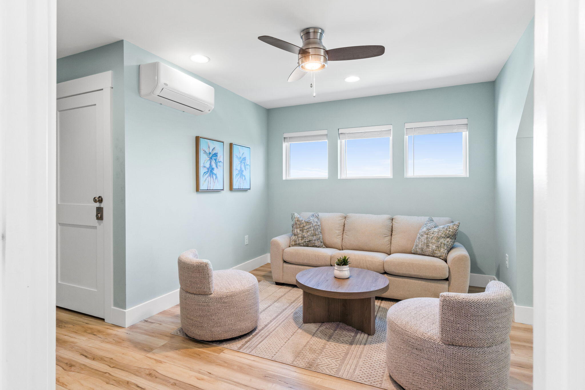 Staged living room with a beige sofa, two rounded chairs, three windows, and pale blue walls.
