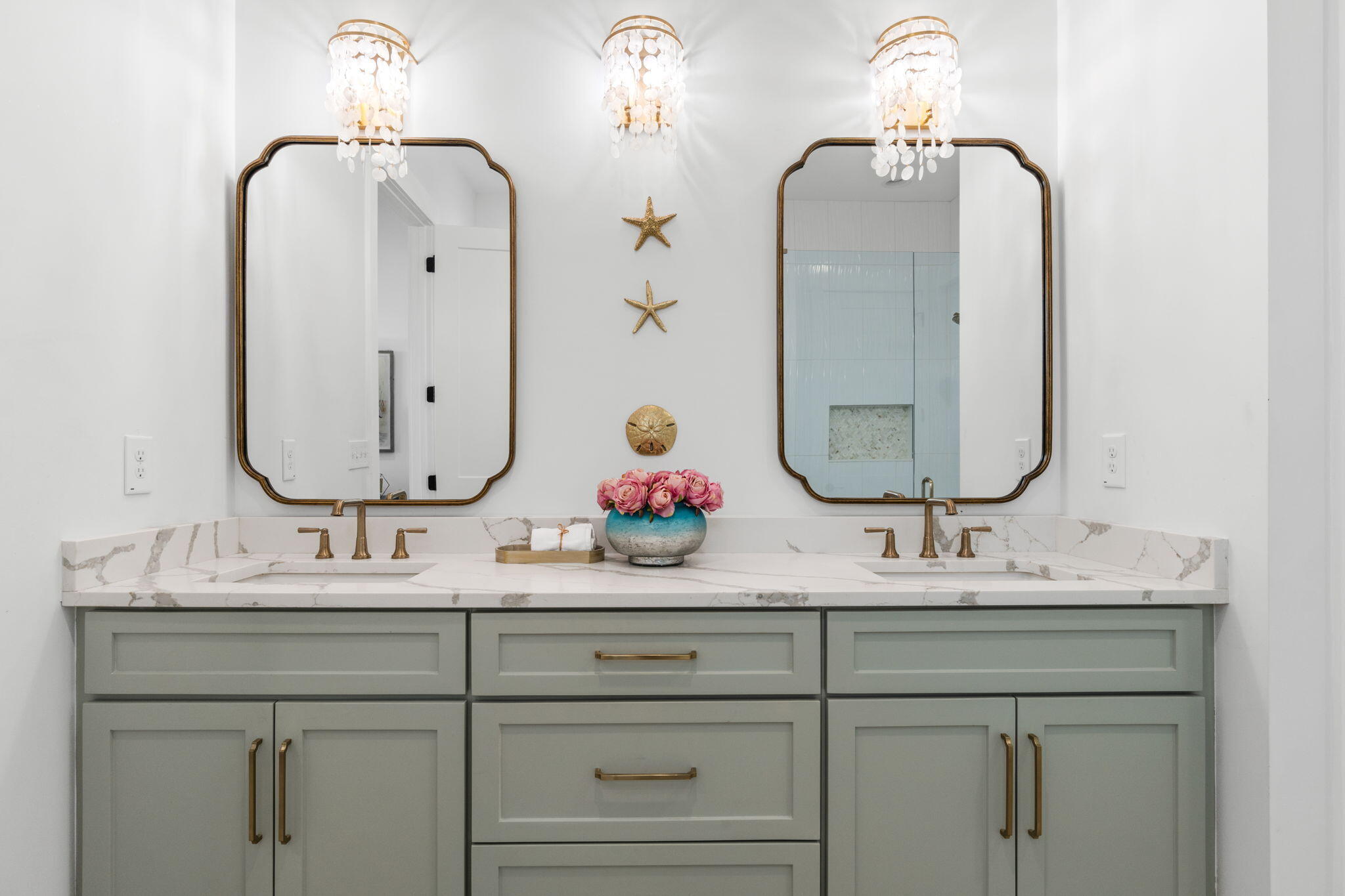 Bathroom double vanity with pale green cabinets, marble countertop, brass fixtures, and coastal decor.