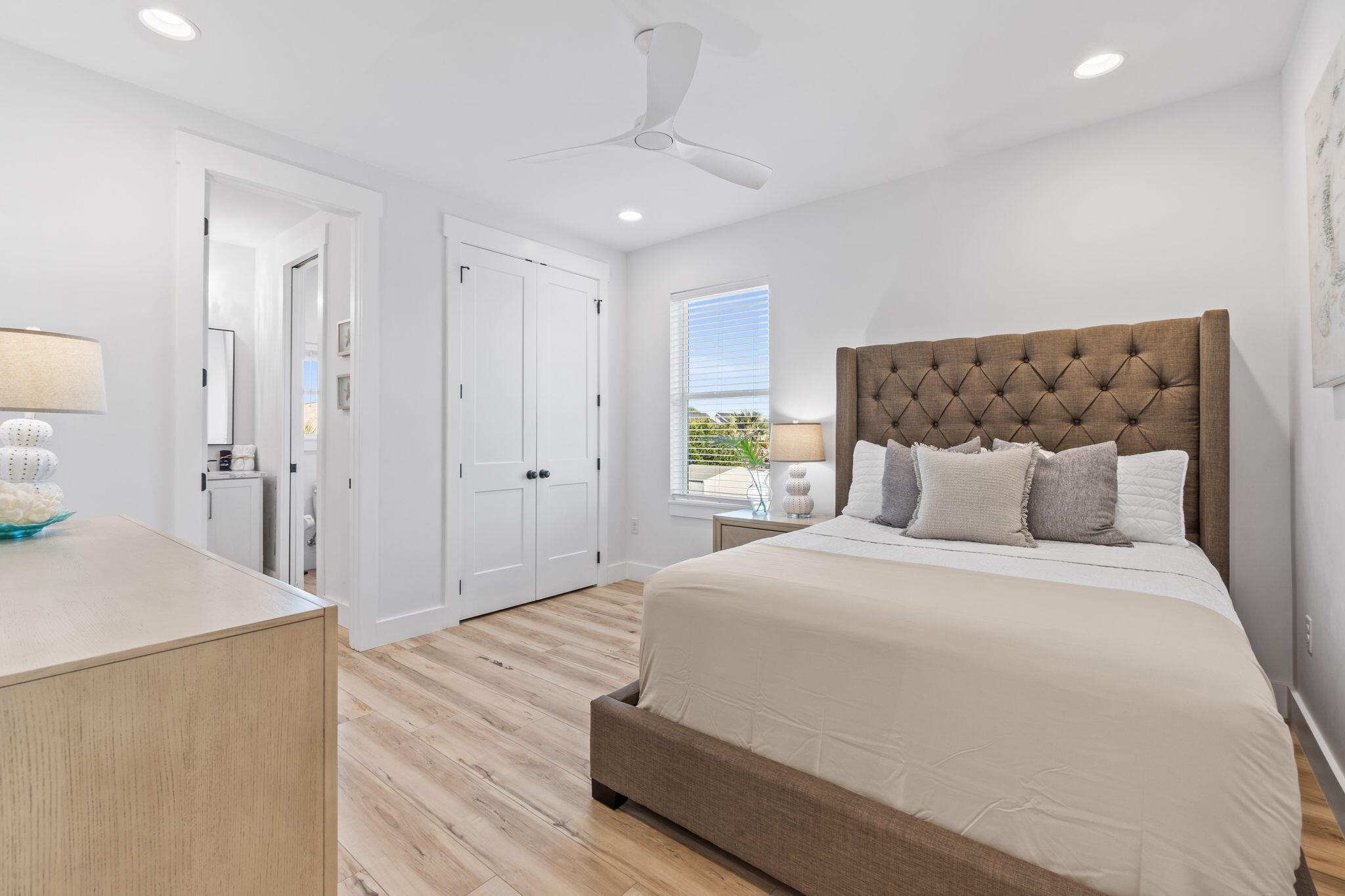 Staged bedroom with a large bed, tufted brown headboard, pale wood floors, and a window with blinds.