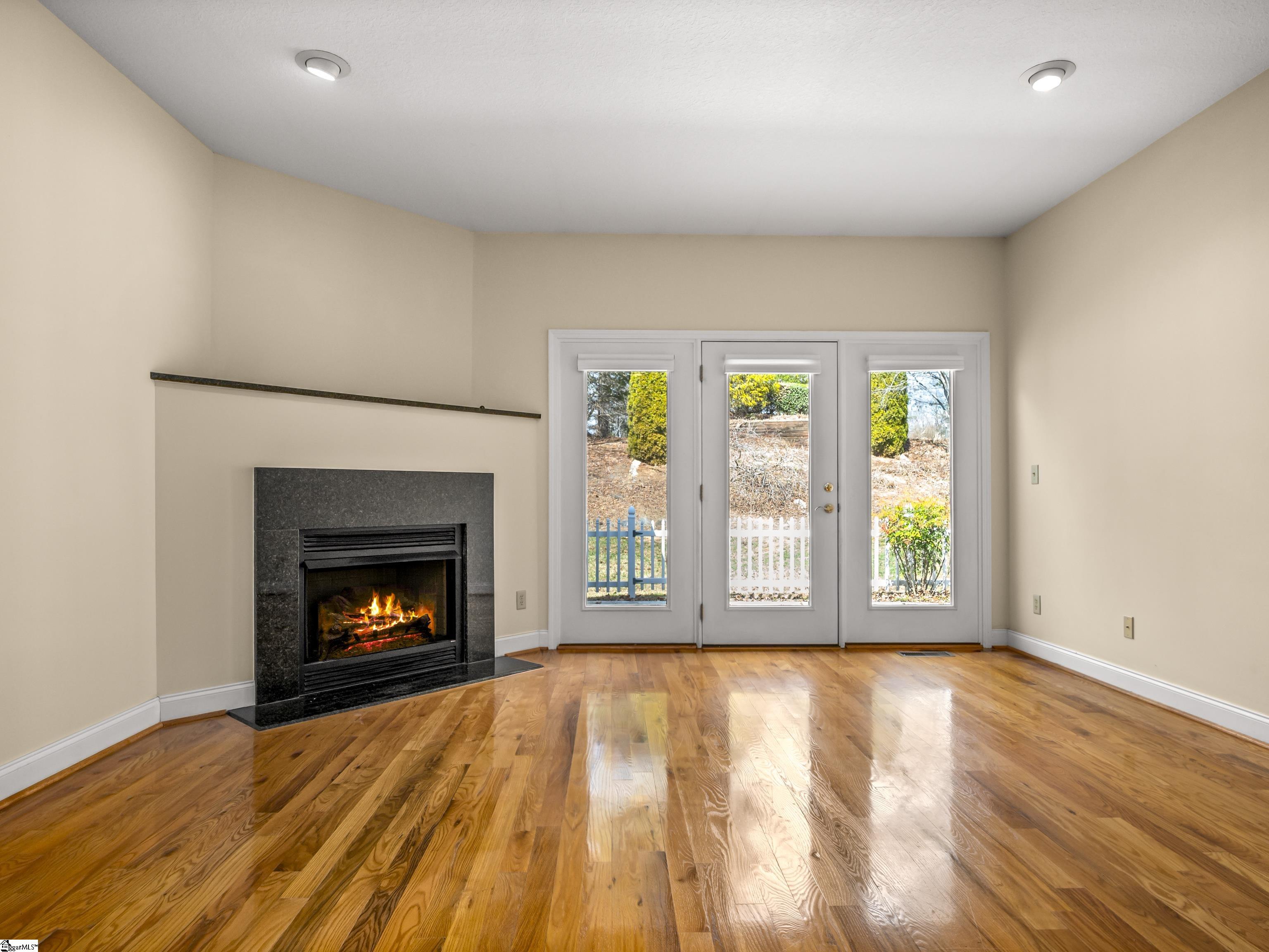 Empty living room with a lit stone fireplace, glossy hardwood floors, and French doors to the yard.