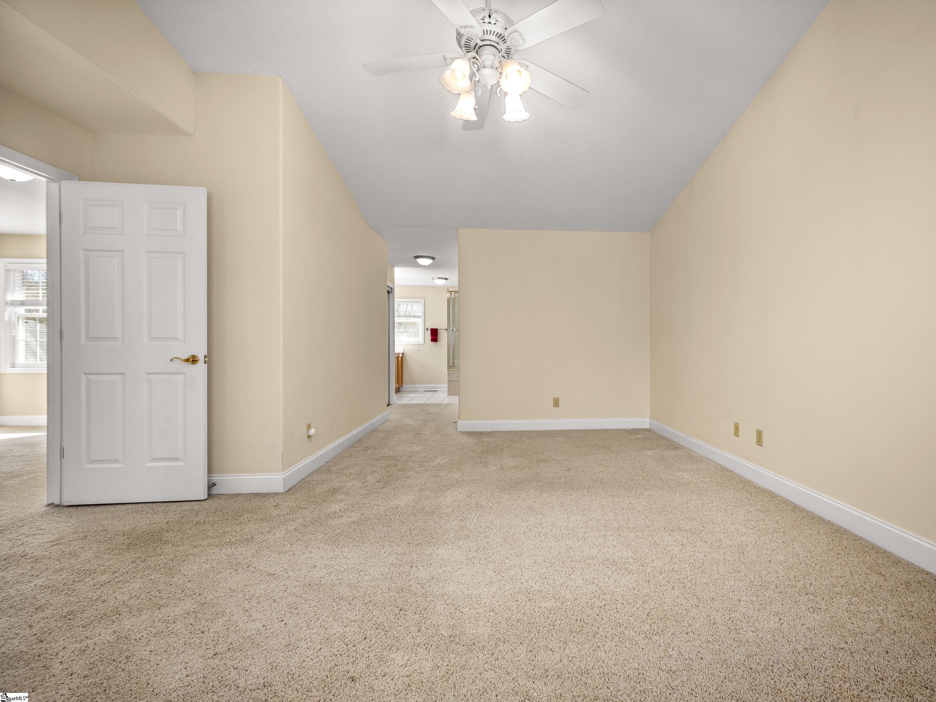 Empty carpeted bedroom with beige walls, a ceiling fan, and an open doorway to a bathroom.