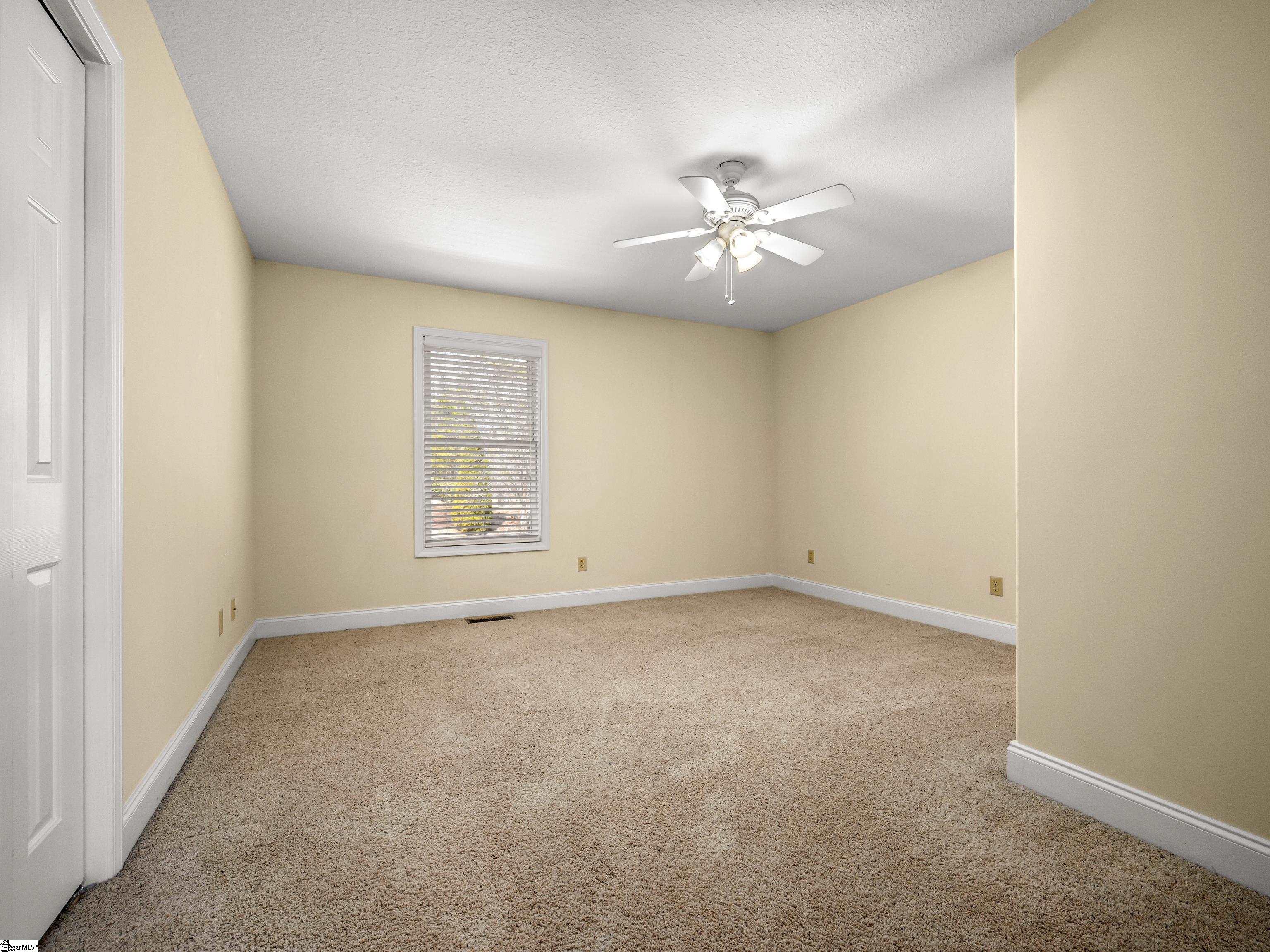 Empty bedroom with beige walls, tan carpet, a window with blinds, and a ceiling fan.