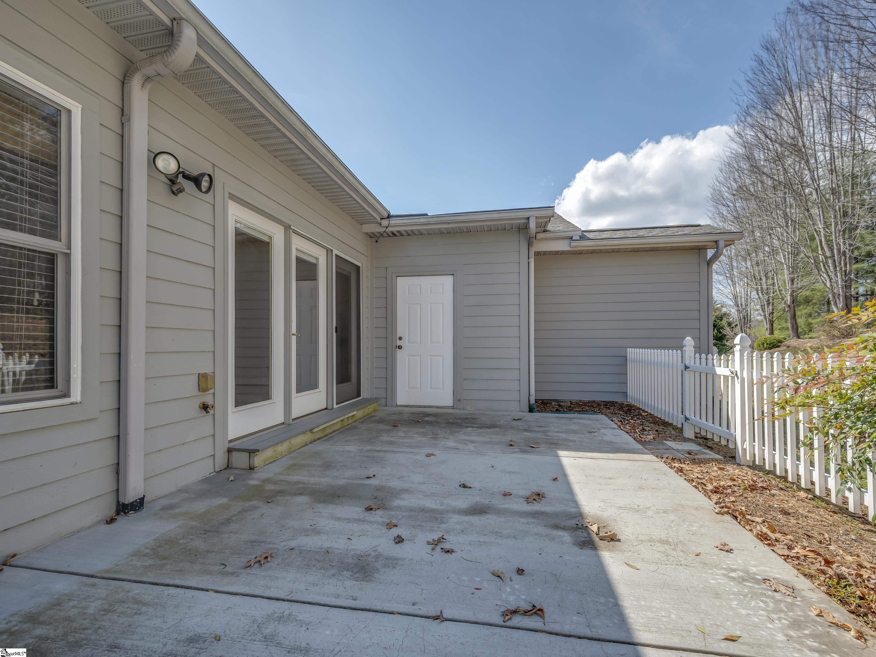 Concrete patio beside a gray-sided home, with glass doors, a white picket fence, and side entrance.
