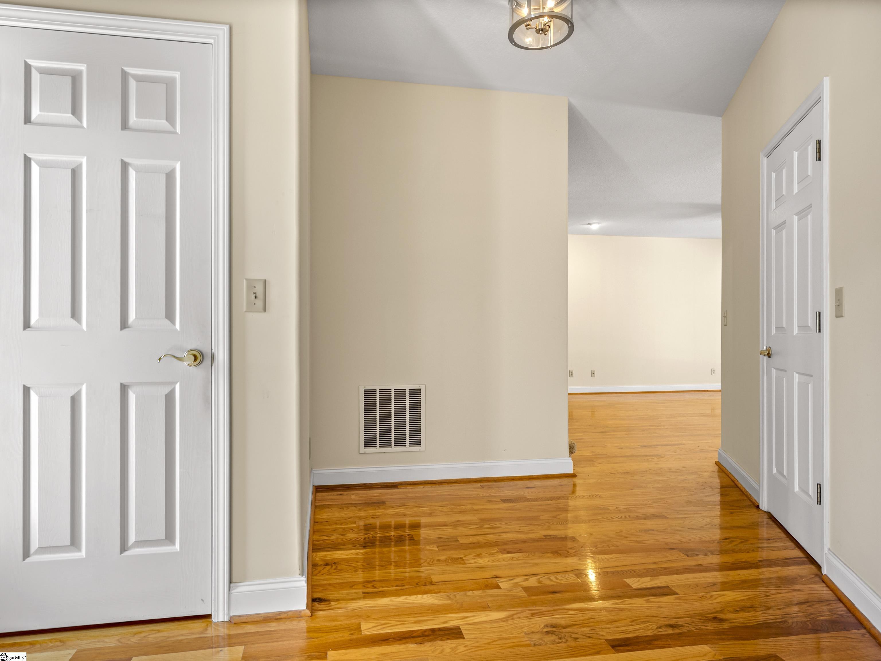 Empty hallway with glossy hardwood floors, cream walls, white paneled doors, and an opening to a larger room.
