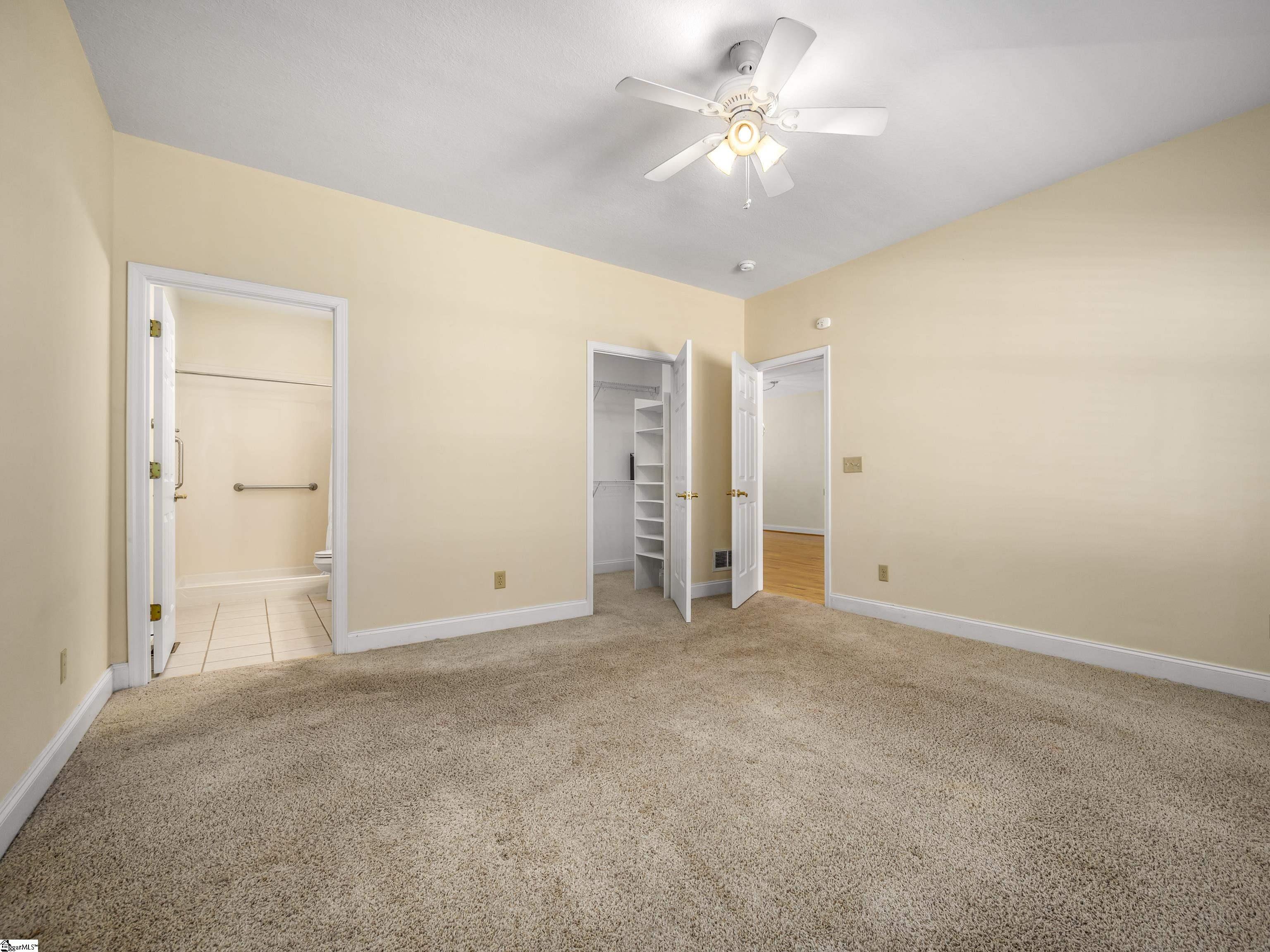 Empty carpeted bedroom with a ceiling fan and open doors to a bathroom and closet.