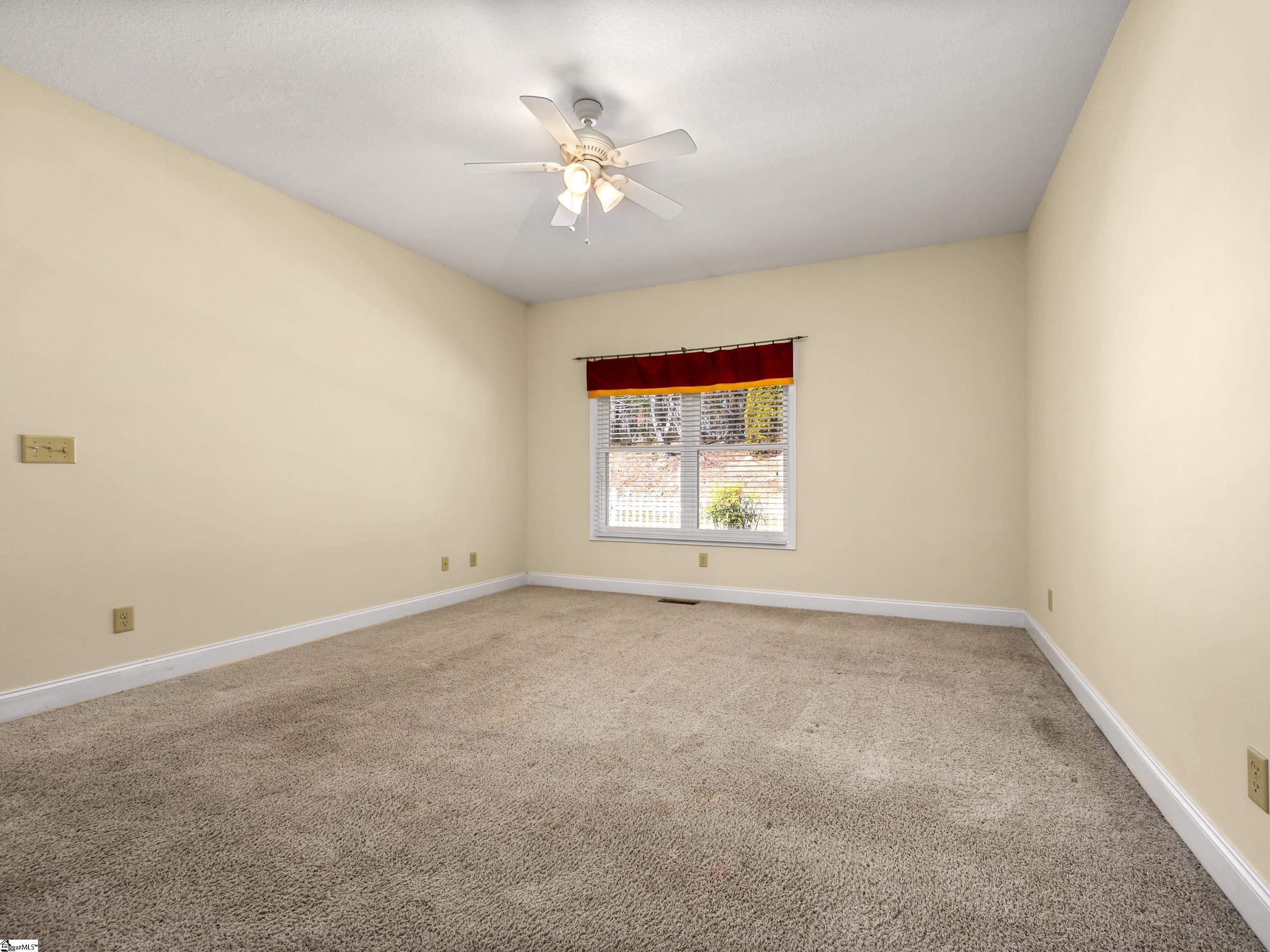 Empty carpeted room with cream walls, a red window valance, and a ceiling fan with lights.