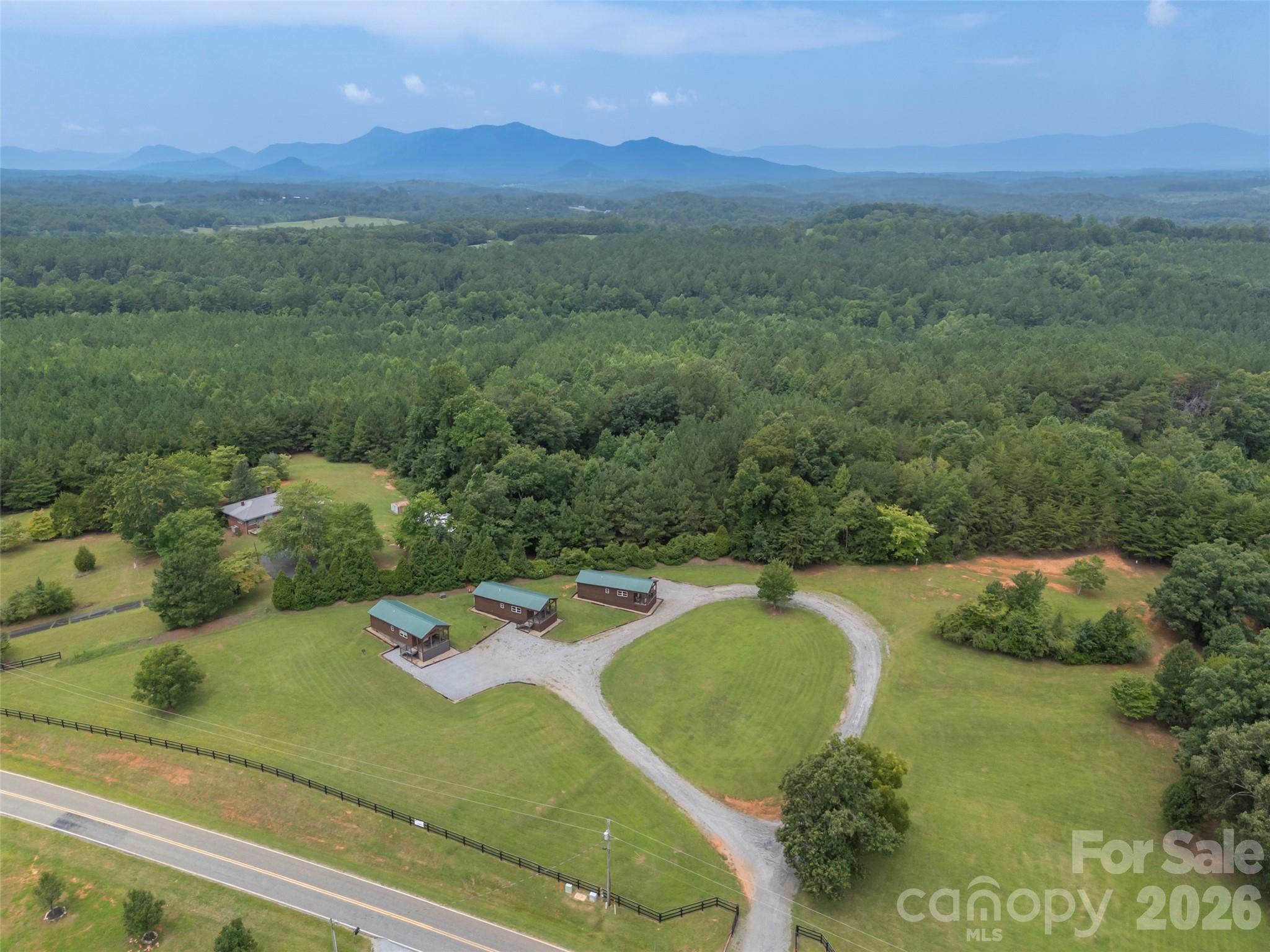 Aerial view of three cabins with a looping gravel driveway, wooded acreage, and distant mountain views.