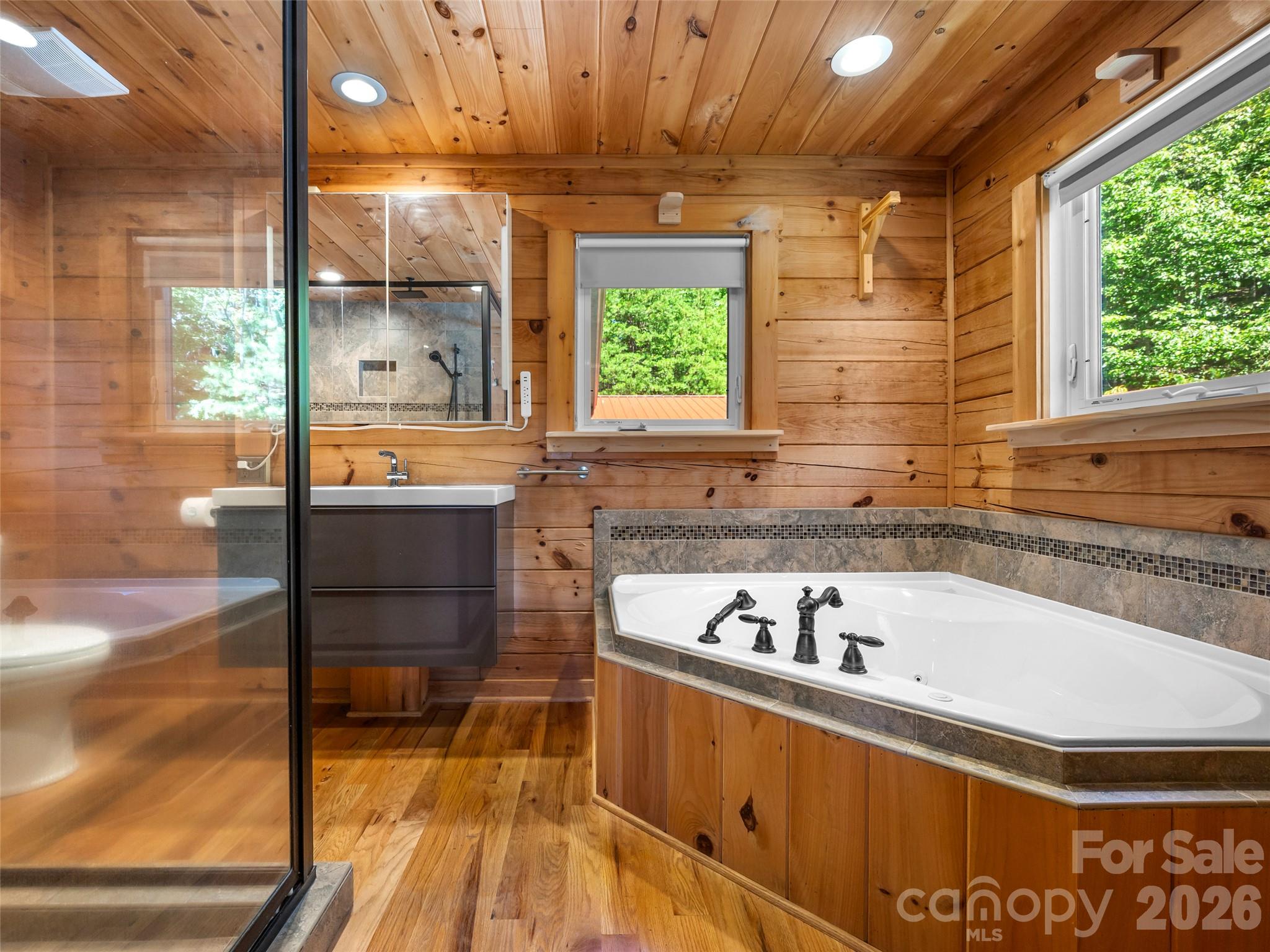 Wood-paneled bathroom with a corner jetted tub, glass shower, and windows overlooking trees.