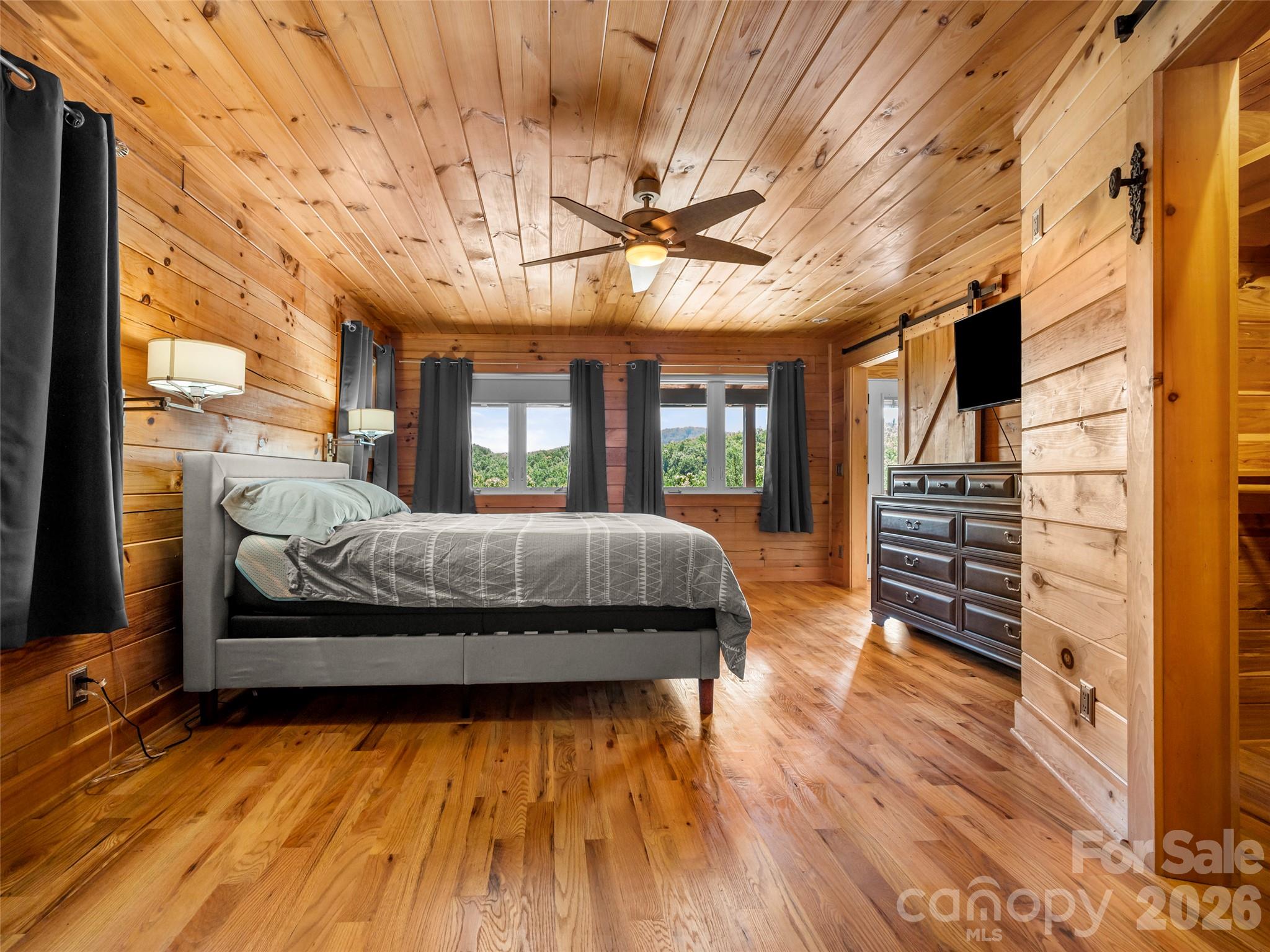 Wood-paneled bedroom with a gray bed, dark curtains, and windows overlooking wooded hills.