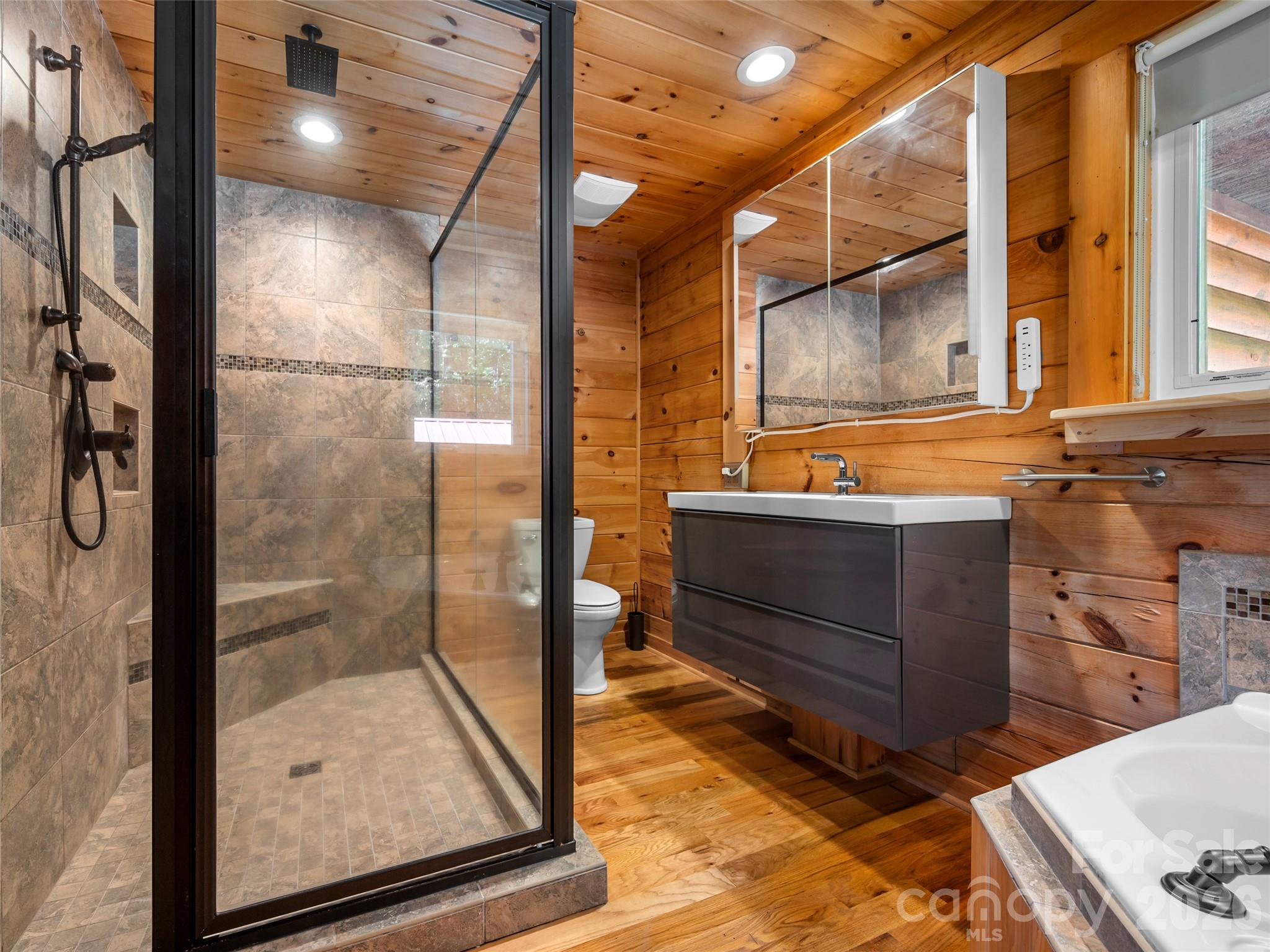 Wood-paneled bathroom with a glass shower, dark floating vanity, and partial bathtub.