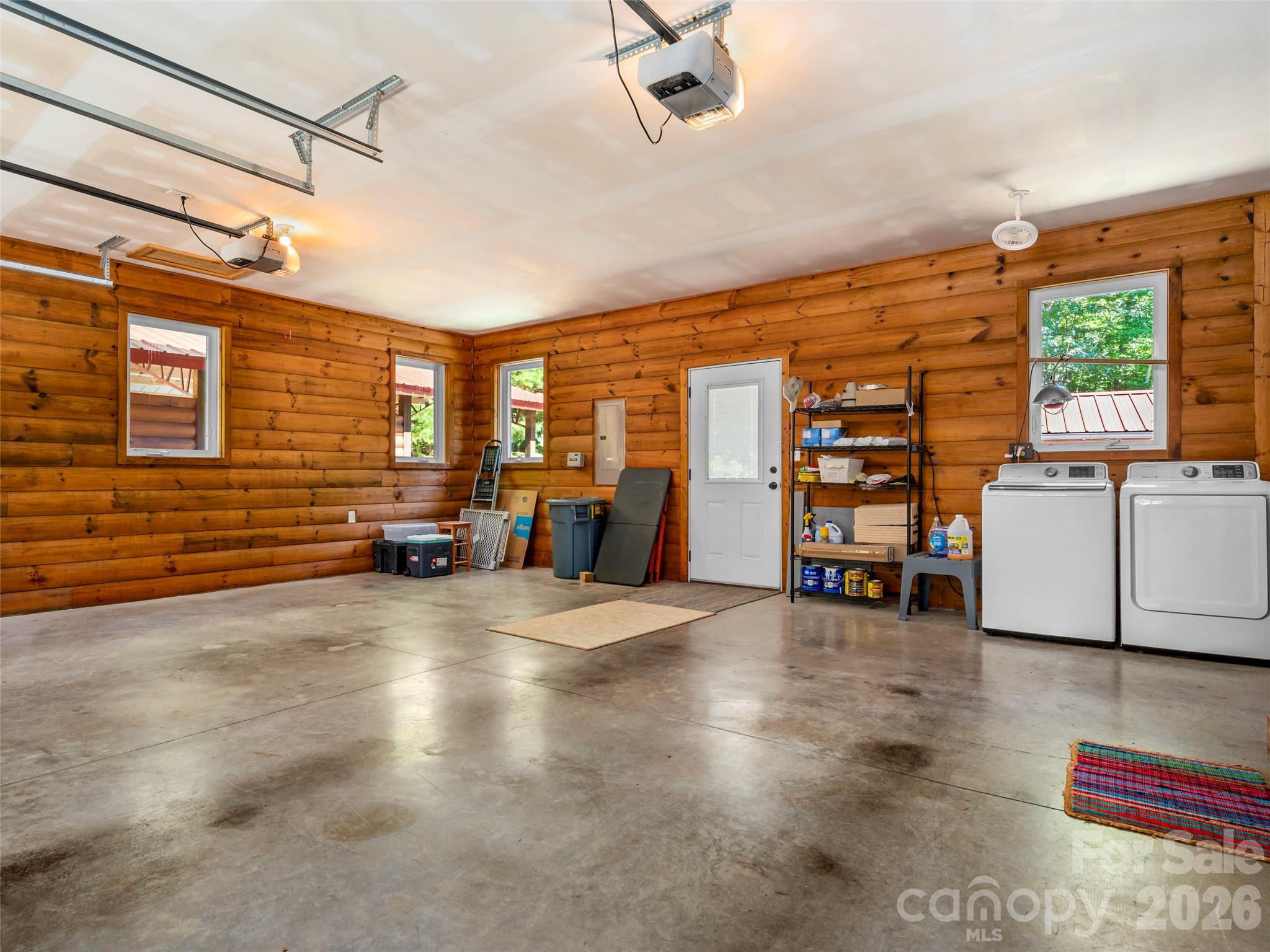 Log-walled garage with concrete flooring, two overhead door openers, storage shelves, and a washer and dryer.