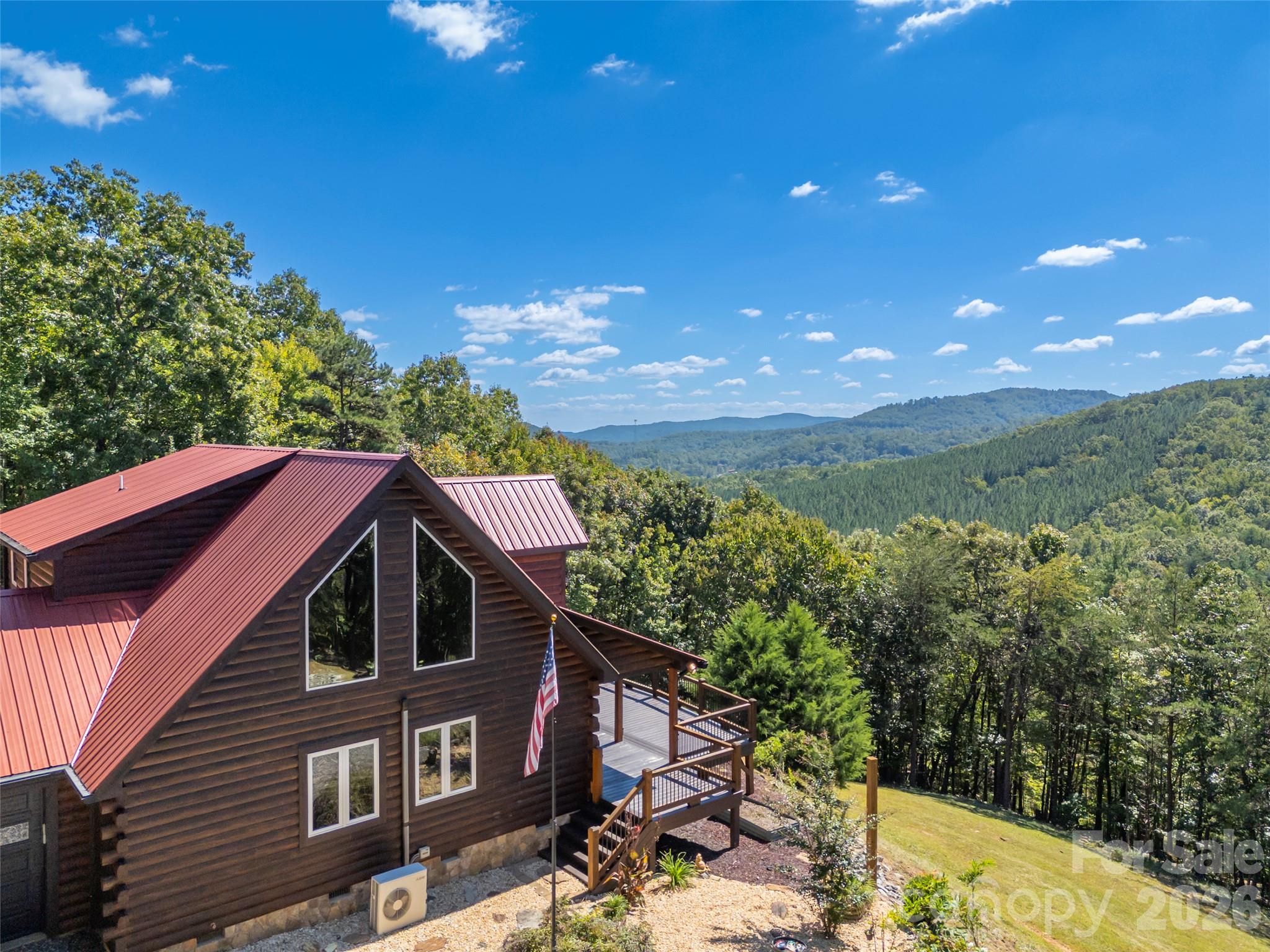 Mountain cabin with a red metal roof and deck overlooking forested hills.