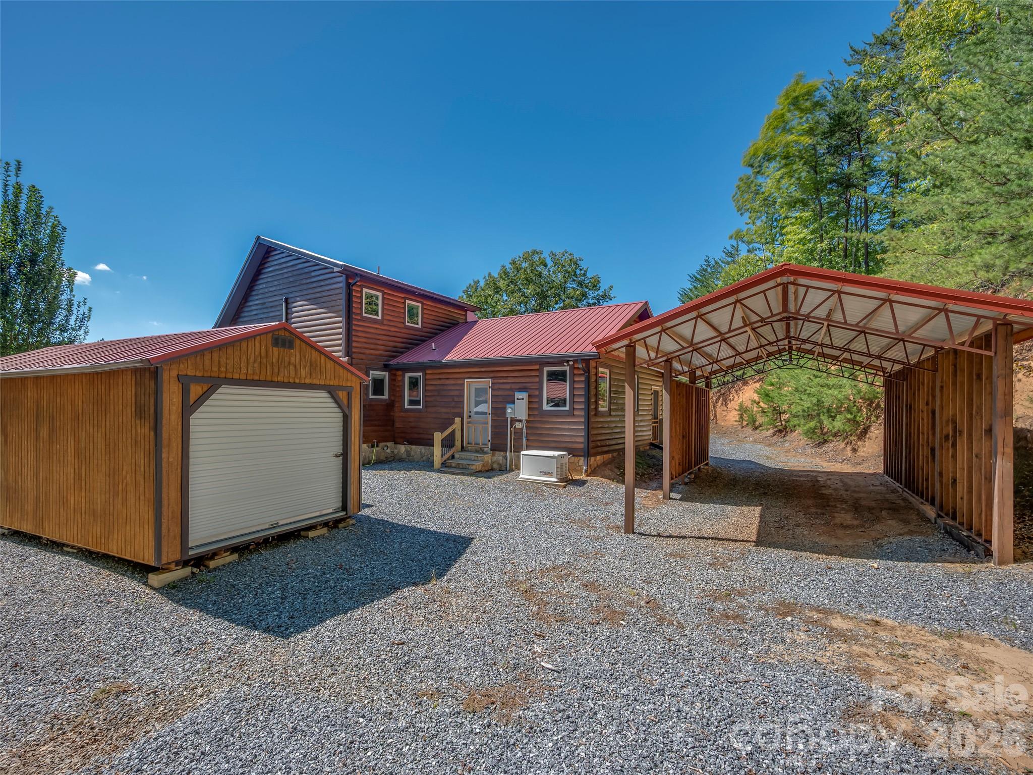 Log-sided home with a detached wooden garage and large metal carport on a gravel driveway.