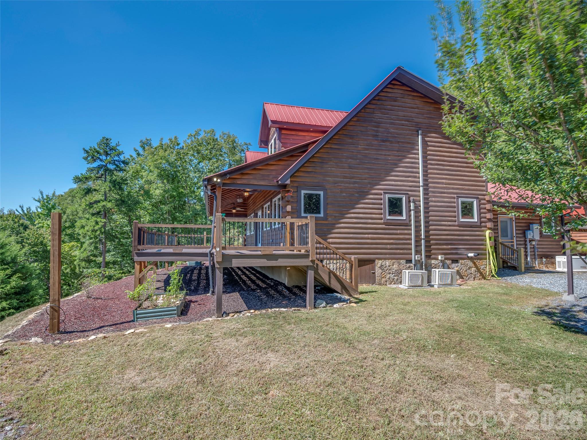 Log-sided home with a red metal roof and raised deck on a grassy, wooded slope.