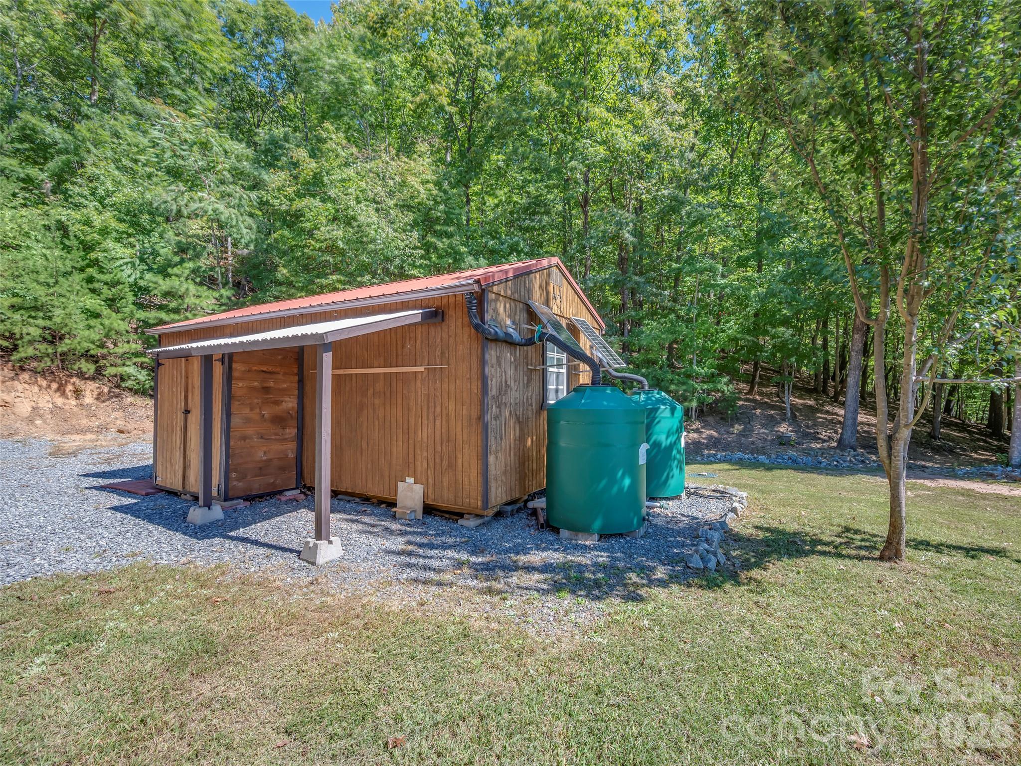 Wooden outbuilding with a red metal roof, solar panels, and two green water tanks in a wooded yard.