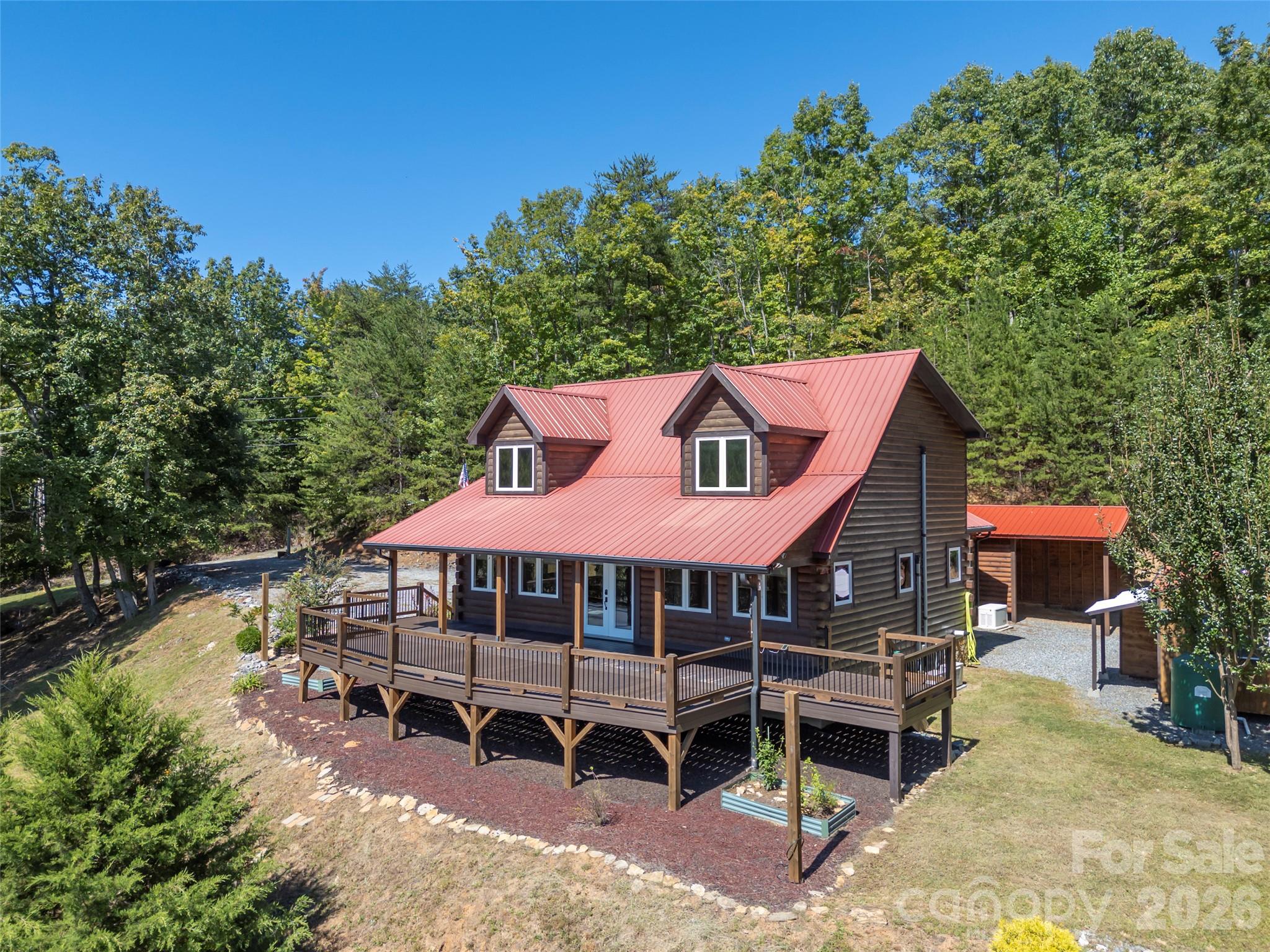 Wood cabin with a red metal roof and raised wraparound deck on a wooded lot.