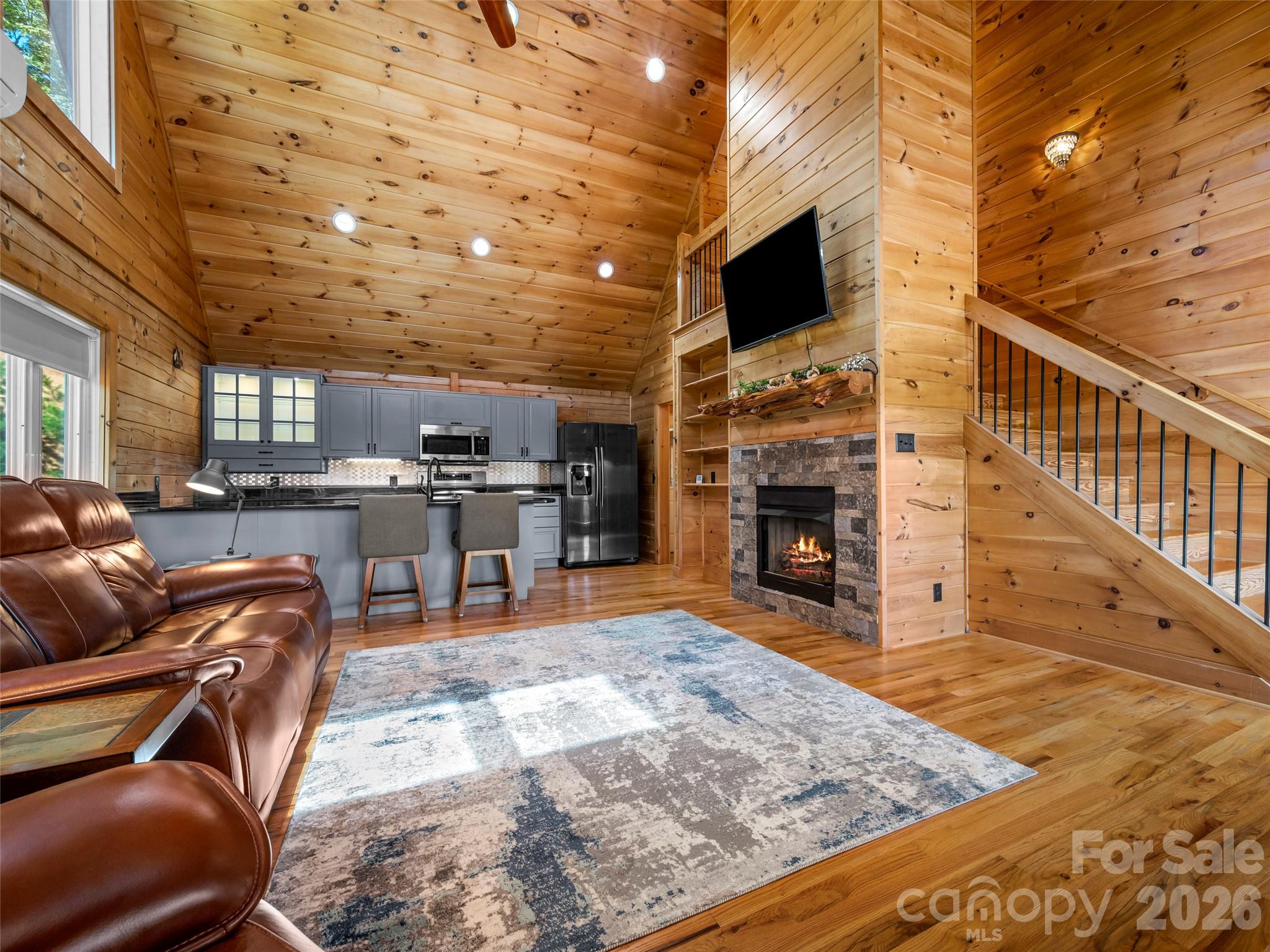 Open-plan living room and kitchen with vaulted pine ceiling, leather sofa, stone fireplace, and staircase.