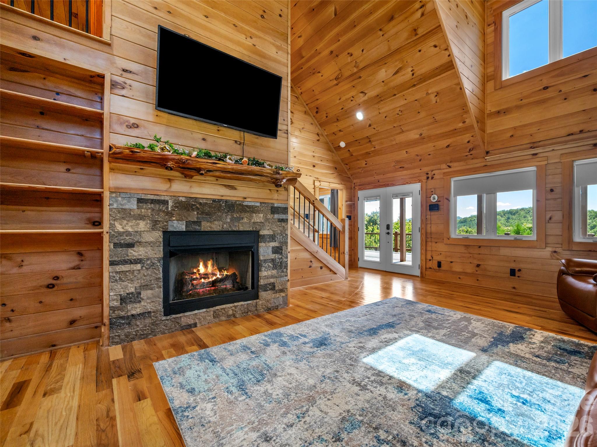 Cabin living room with a stone fireplace, vaulted wood ceiling, and windows overlooking wooded hills.