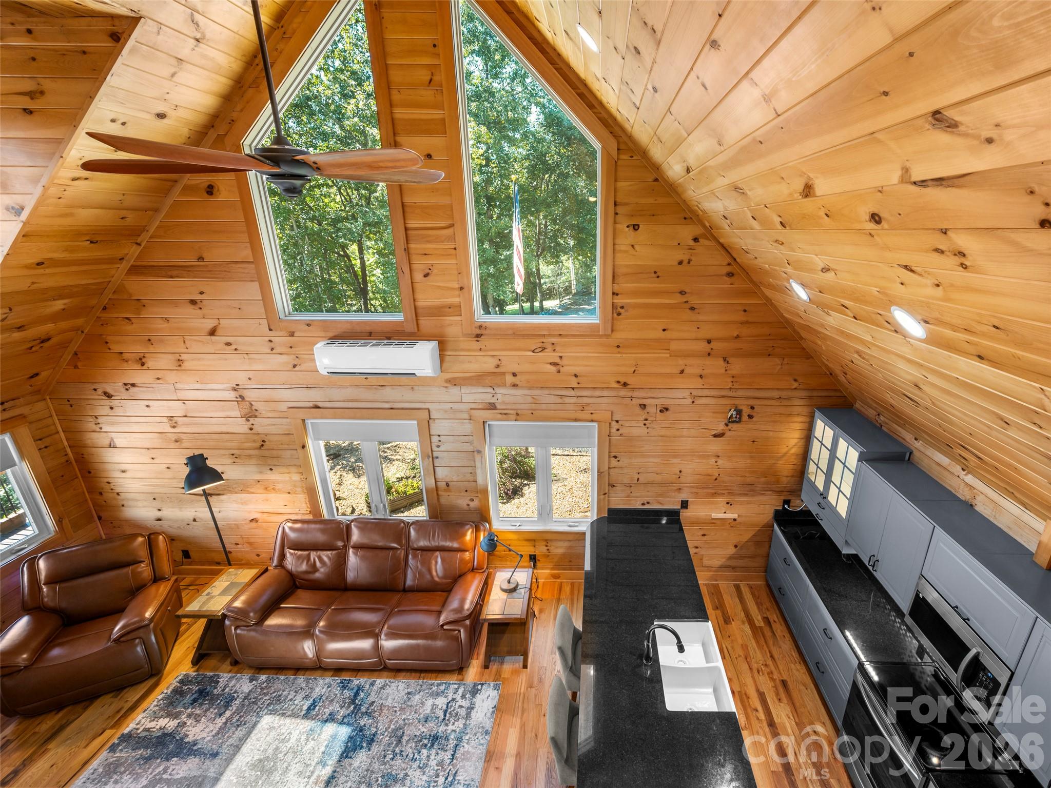 Vaulted wood-paneled living room and kitchen with tall windows, leather seating, and gray cabinets.