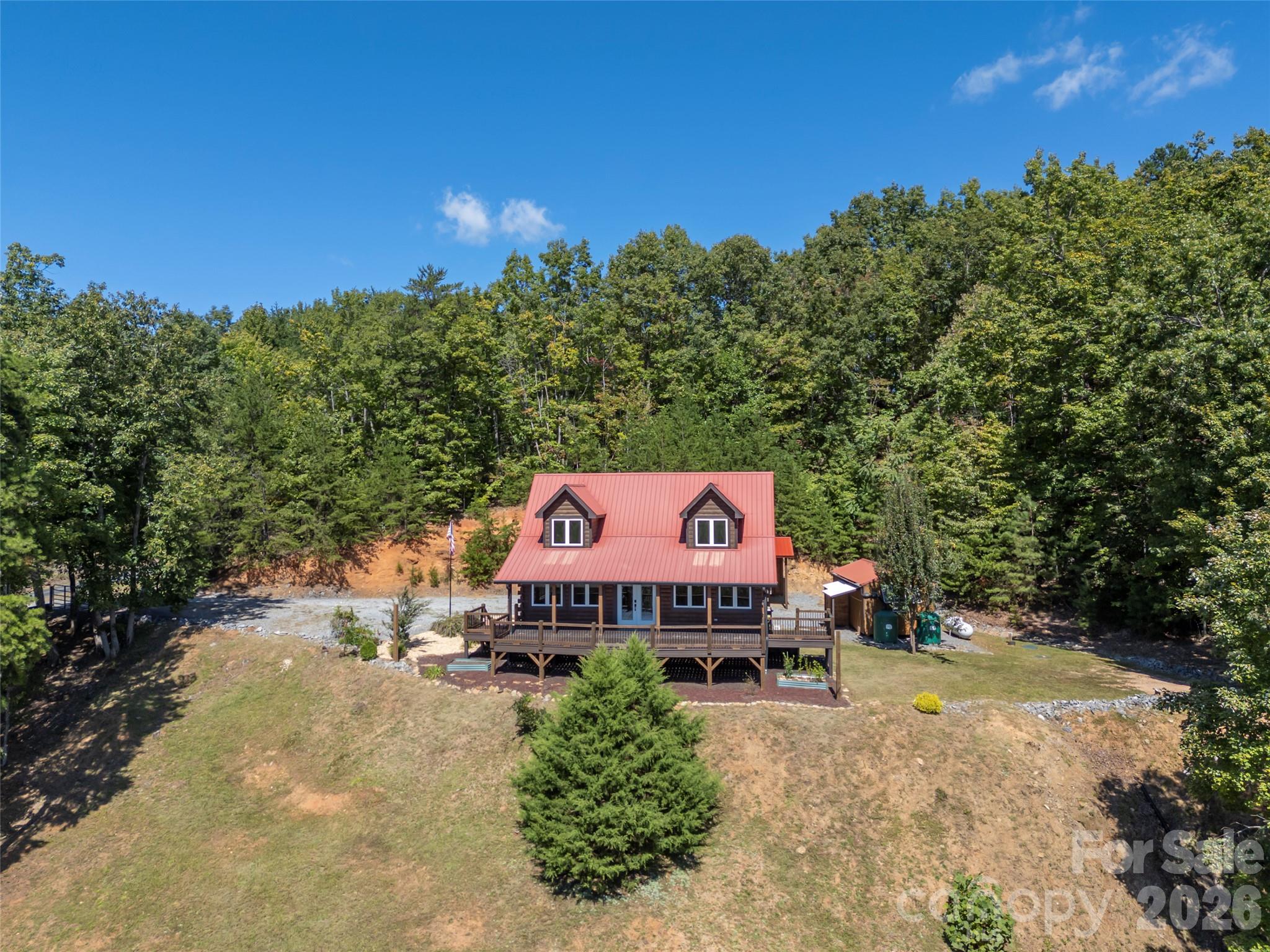 Wooded home with a red metal roof, two dormers, and a wraparound porch, viewed from above.
