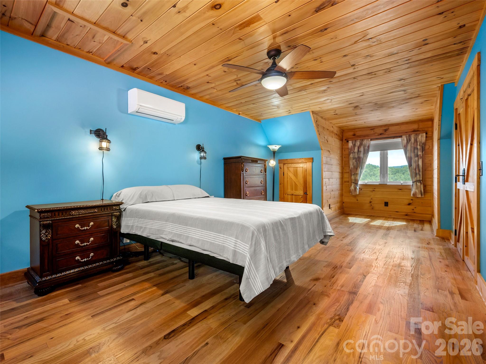 Bedroom with blue walls, vaulted pine ceiling, hardwood floors, and a window overlooking wooded hills.