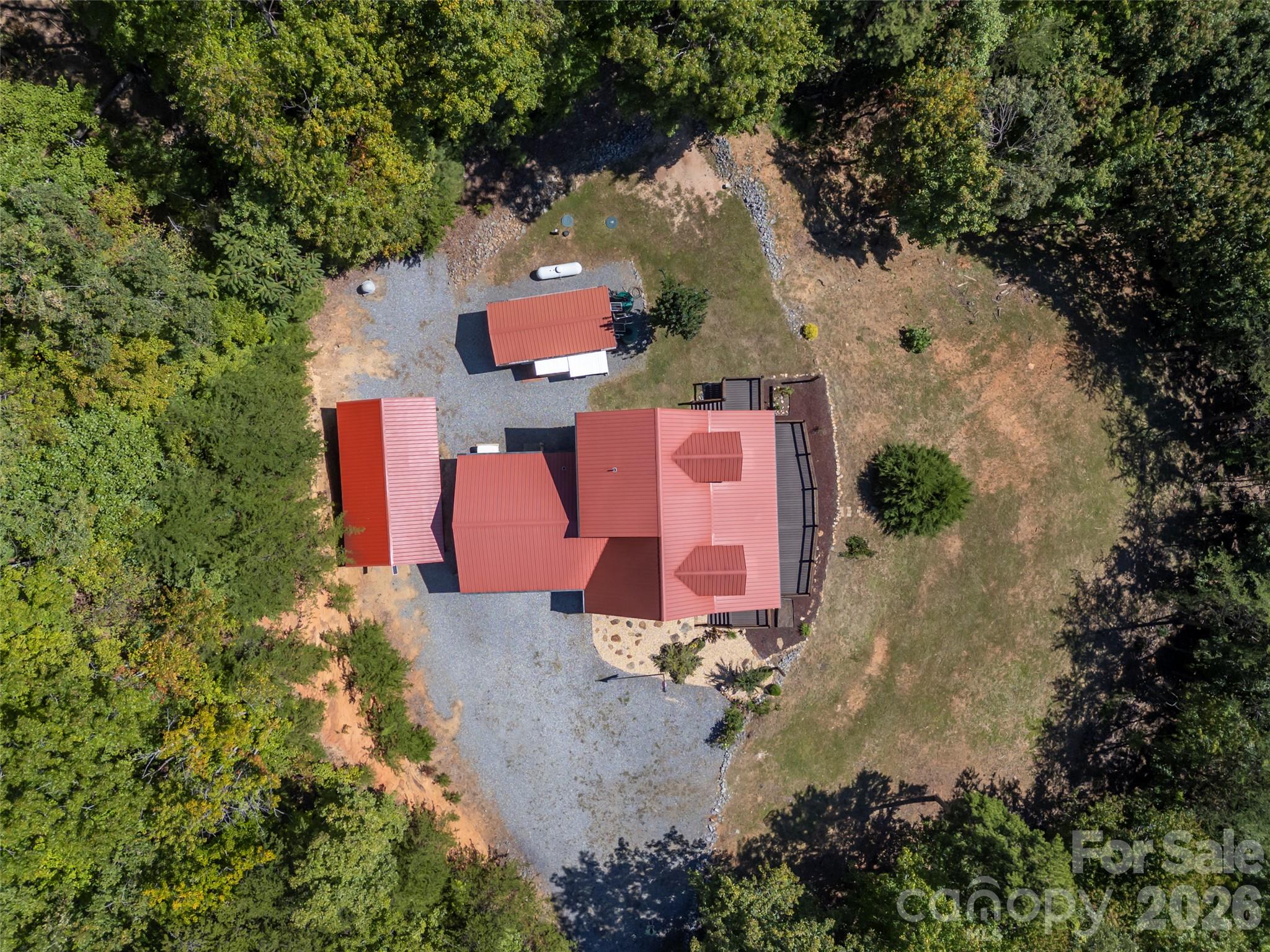 Aerial view of a red-roofed home and outbuilding in a wooded clearing with a broad gravel driveway.