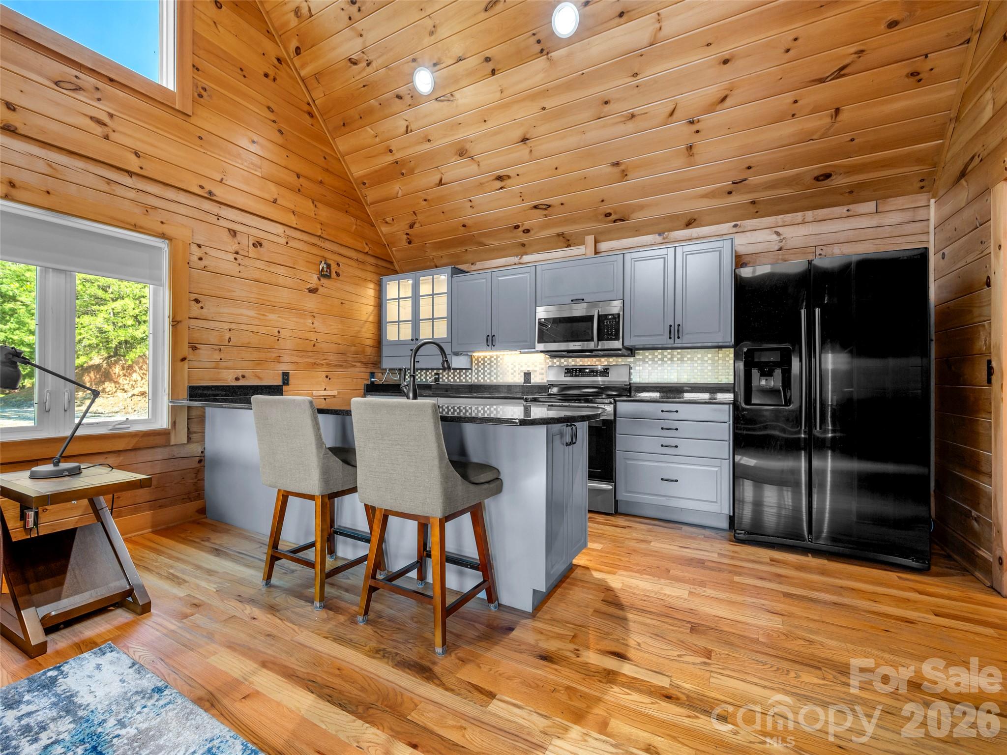 Kitchen with gray cabinets, black appliances, island seating, and vaulted wood-plank ceiling.
