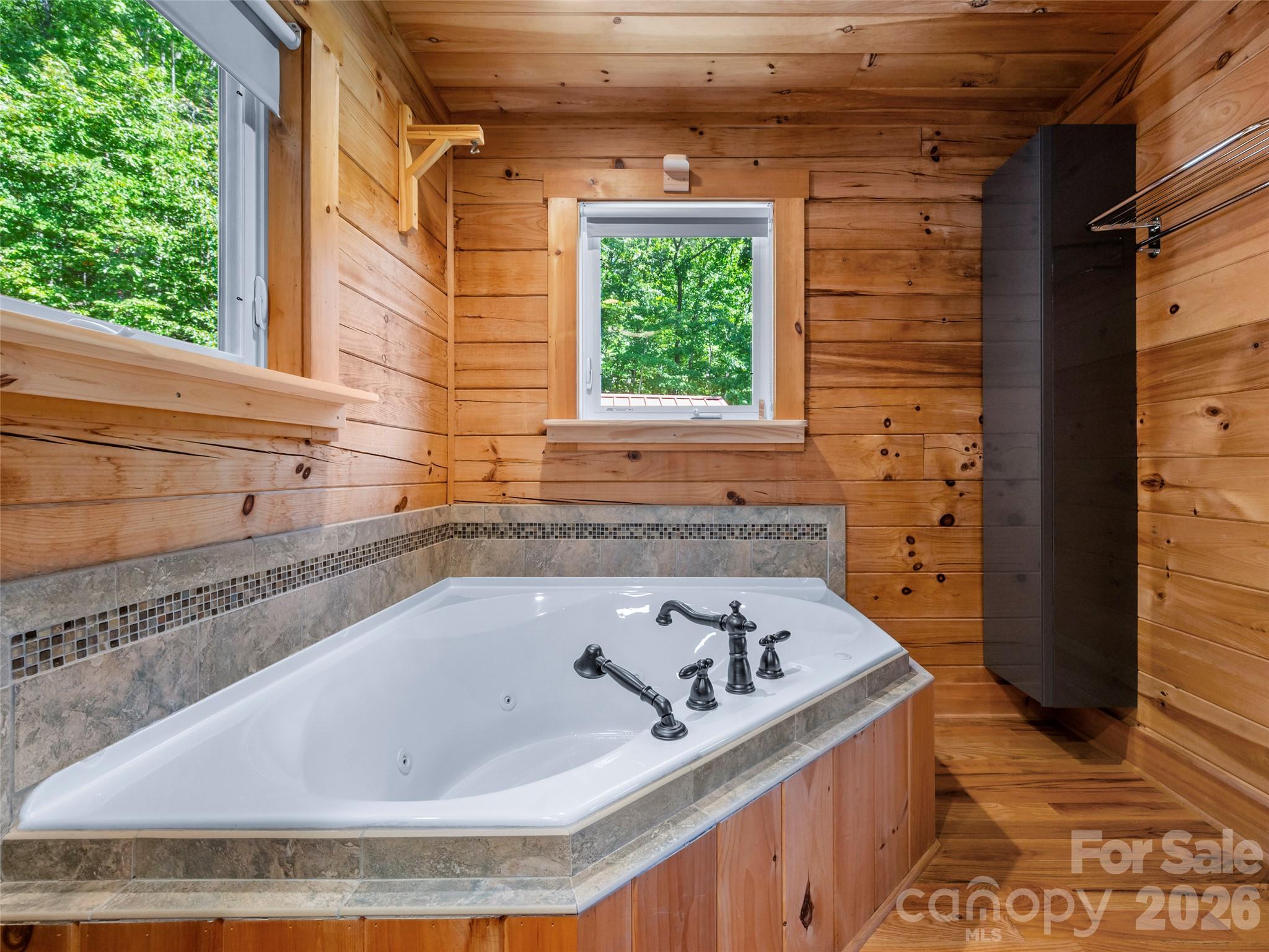 Jetted soaking tub in a wood-paneled bathroom, with stone tile surround and windows overlooking trees.