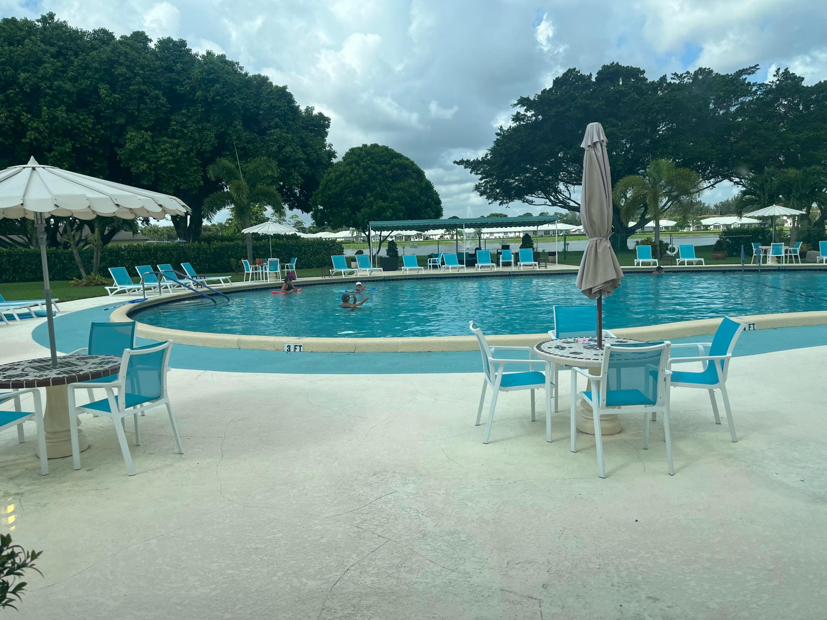 Community swimming pool with turquoise water, blue lounge chairs, patio tables, and large shade trees.