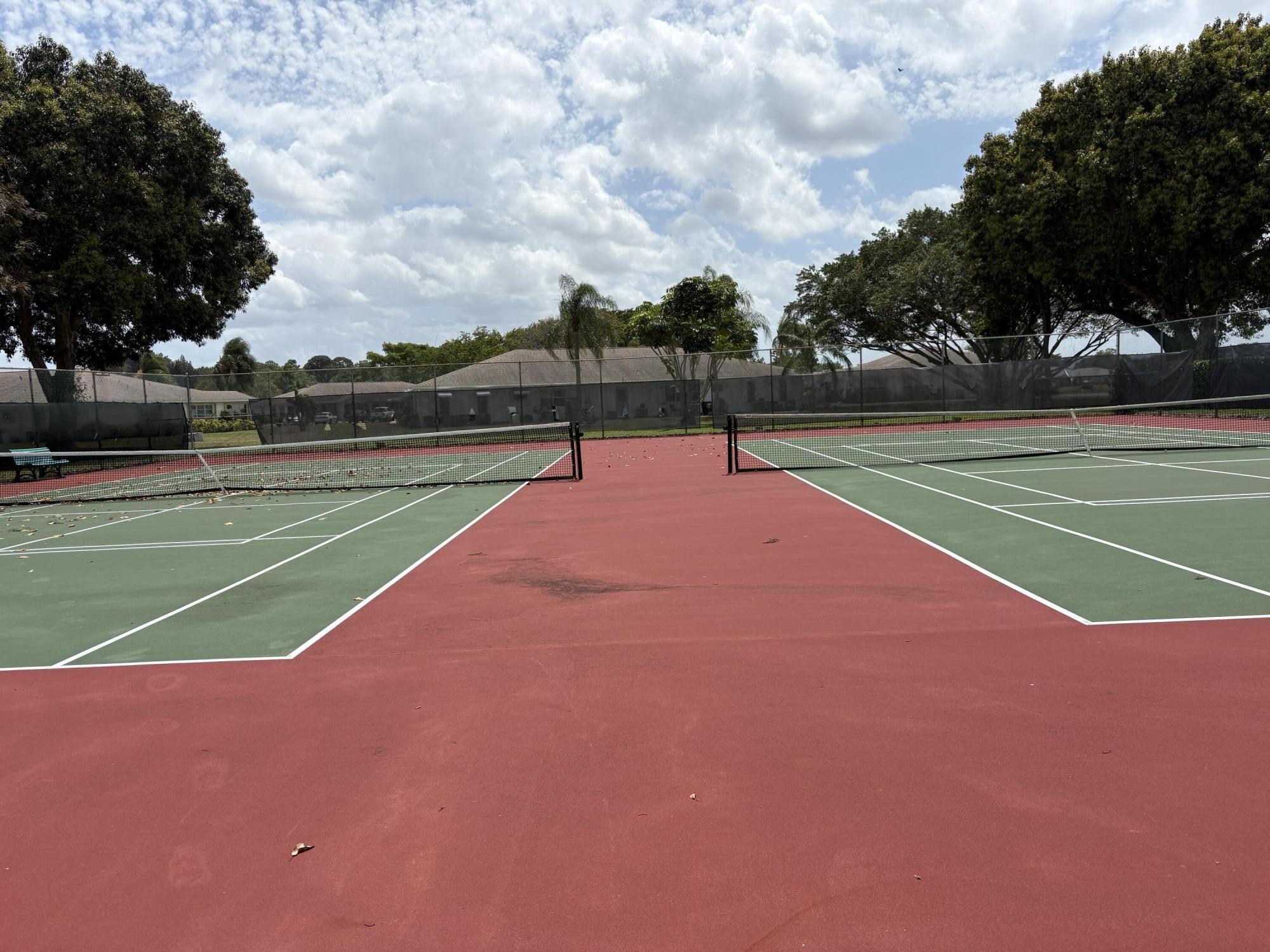 Outdoor tennis courts with green playing surfaces, red surrounds, and mature trees.