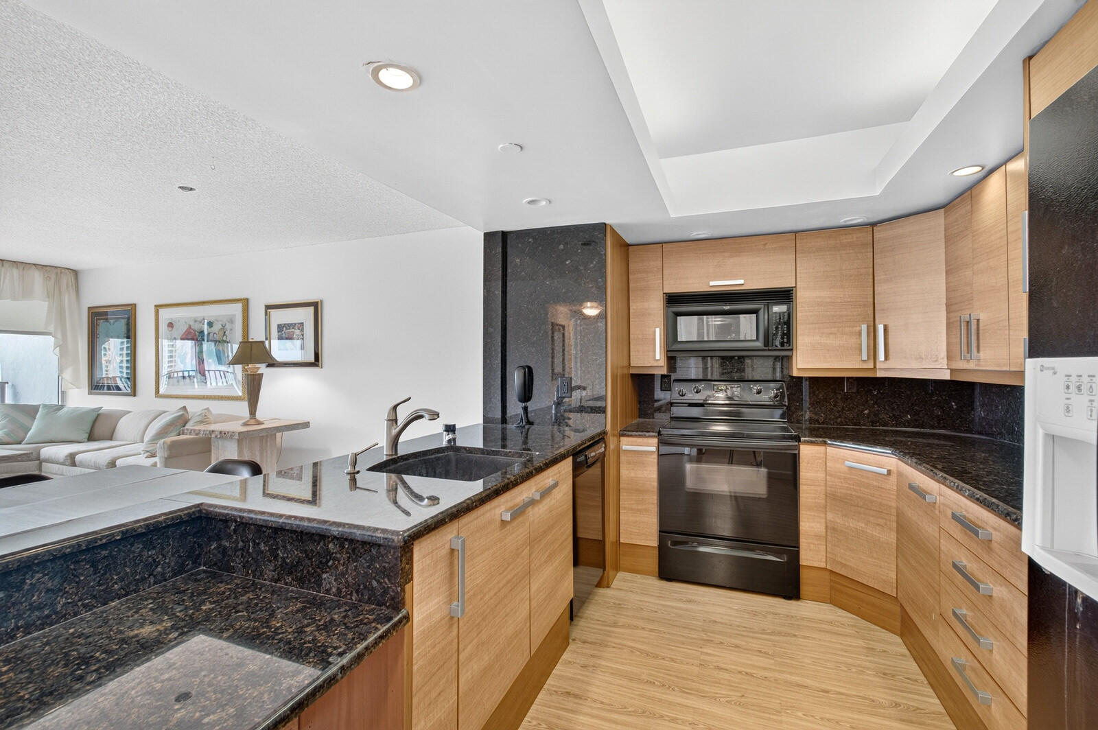 Condominium kitchen with light wood cabinets, dark stone counters, and stainless steel appliances beside the living area.