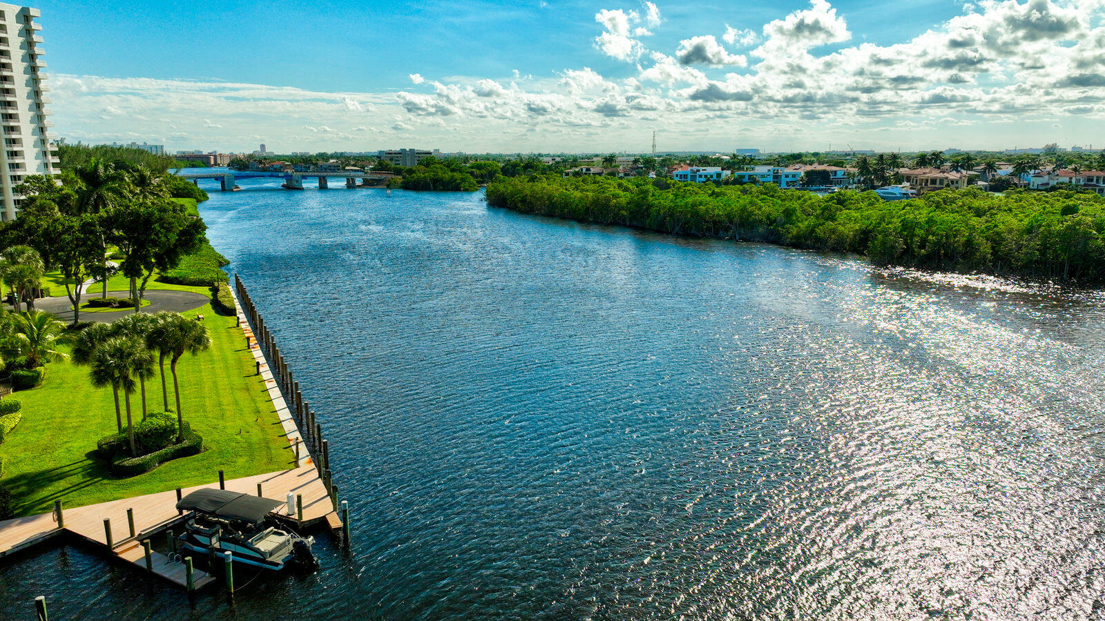 Waterway view with a tree-lined shore, bridge, landscaped lawn, and private dock with a boat.