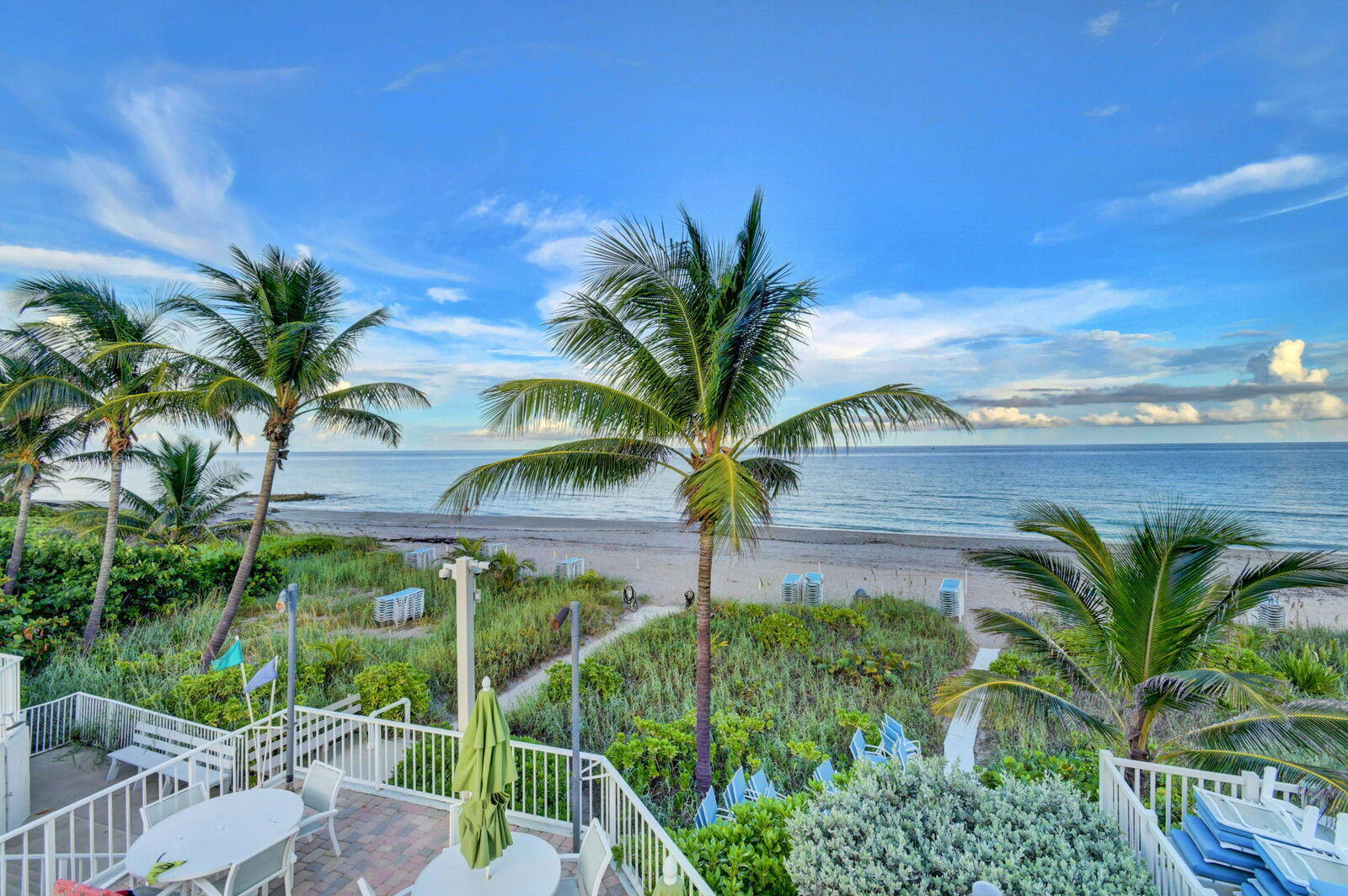 Oceanfront terrace with patio seating, palm trees, sandy beach, and calm water.
