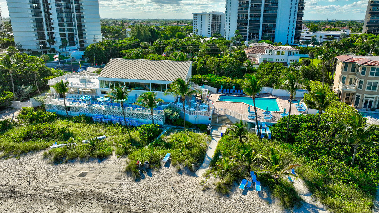 Beachfront condominium grounds with a pool, clubhouse, palm trees, and sandy beach.